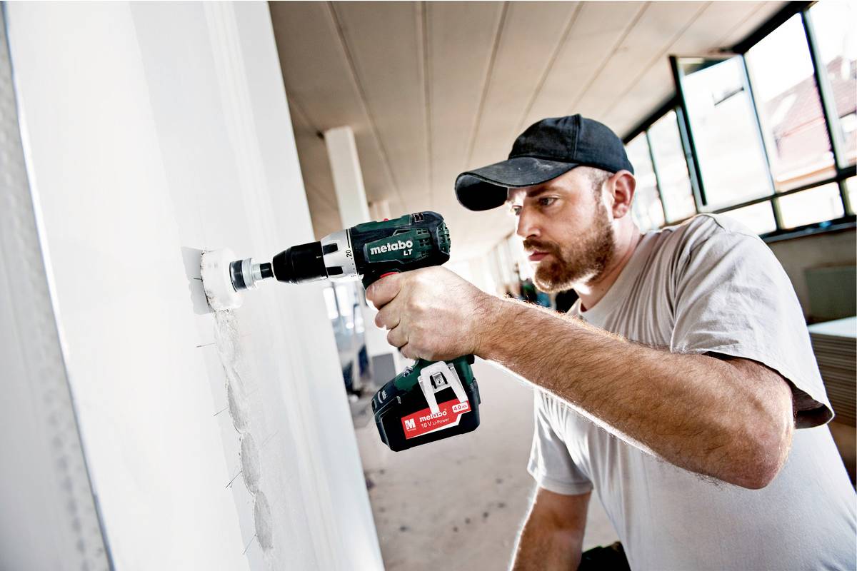 Ein Handwerker bohrt ein großes Loch in die weiße Wand eines hellen Raumes mit einer grünen Bohrmaschine.
