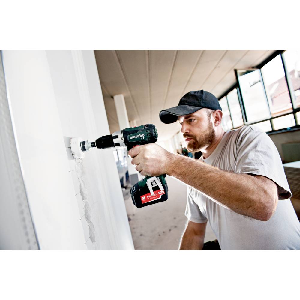Ein Mann in einem weißen T-Shirt bohrt mit einer kabellosen Bohrmaschine ein Loch in eine weiße Wand in einem hellen Raum.