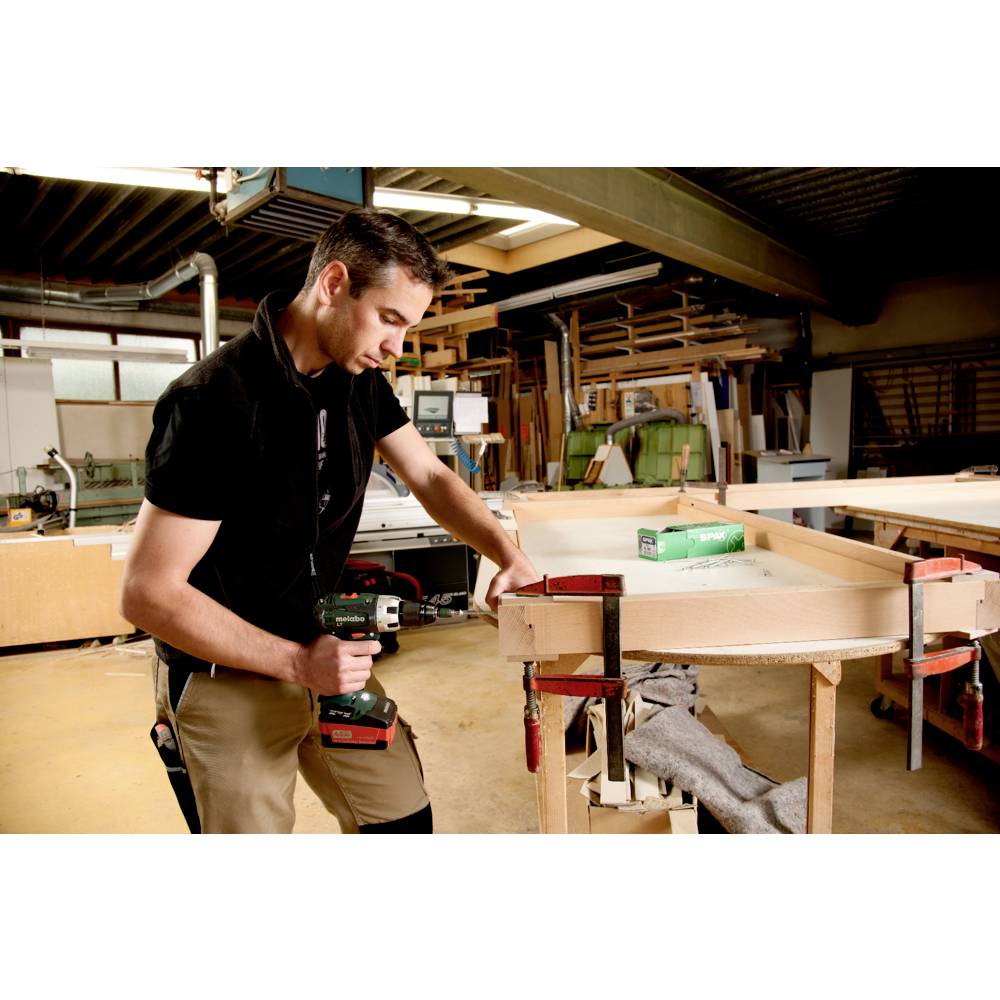 Ein Mann in einer Werkstatt bearbeitet mit einem Akku-Bohrschrauber ein Holzbrett, umgeben von Werkzeugen und Materialien.