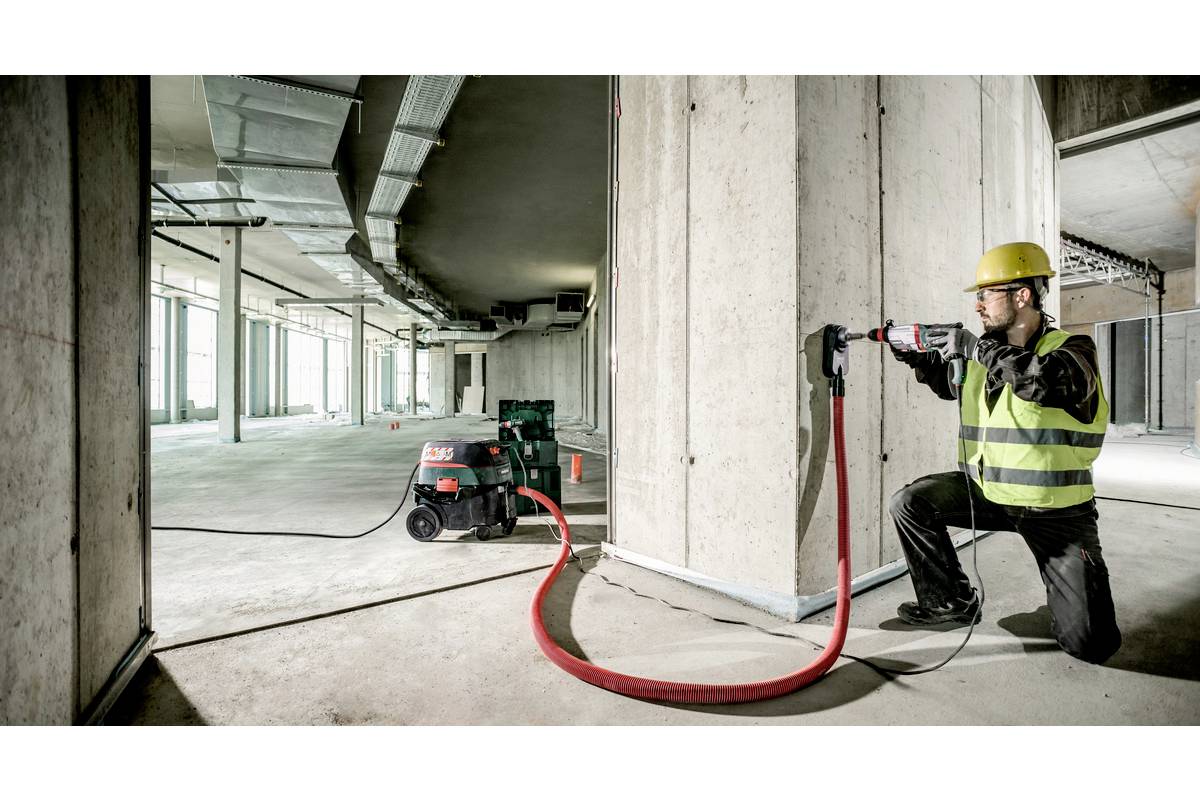 Ein Bauarbeiter in Schutzkleidung nutzt ein Elektrowerkzeug, um ein Loch in eine Betonwand zu bohren. Eine Maschine zur Staubabsaugung ist angeschlossen.