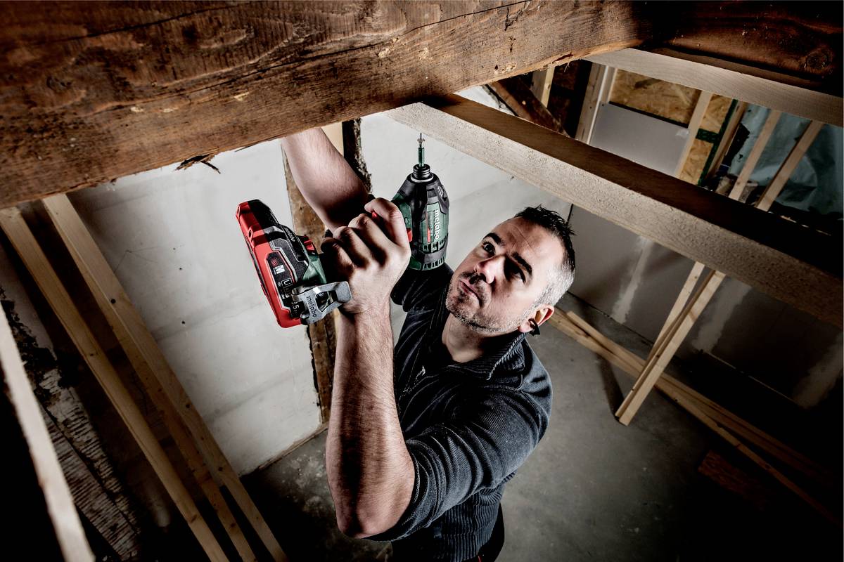 Ein Mann benutzt eine Bohrmaschine, um an einem Holzrahmen zu arbeiten. Er trägt ein dunkelgraues Hemd und befindet sich in einem halbfertigen Raum.