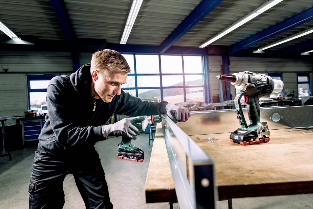 Ein Mann arbeitet in einer Werkstatt und benutzt eine handgehaltene Bohrmaschine, um ein Metallstück an einer Werkbank zu befestigen.