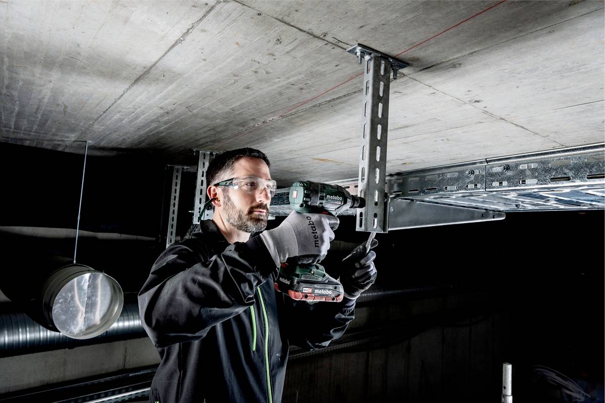 Ein Mann mit Schutzbrille und Handschuhen befestigt eine Metallkonstruktion an der Decke mit einer Bohrmaschine in industrieller Umgebung.