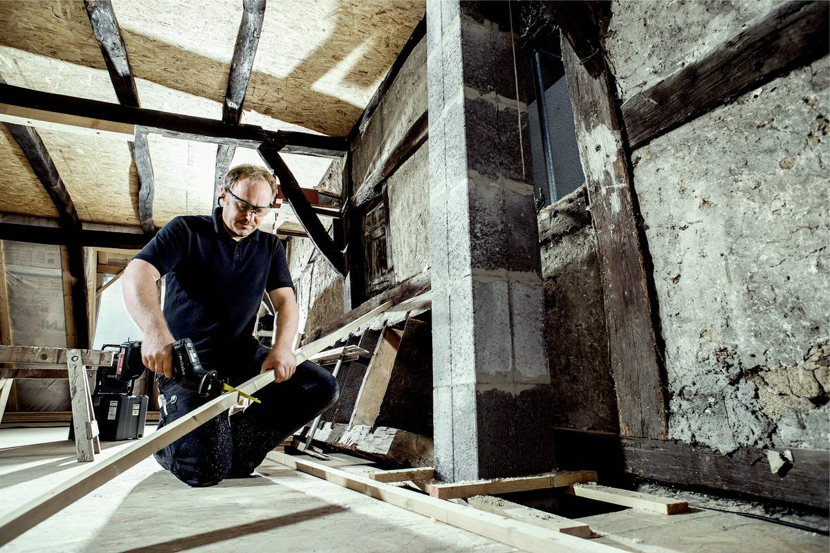 Ein Handwerker kniet in einem Gebäude und nutzt eine Bohrmaschine, um ein Holzbrett zu befestigen. Die Umgebung zeigt eine Baustelle mit Holzbalken und unverputzten Wänden.