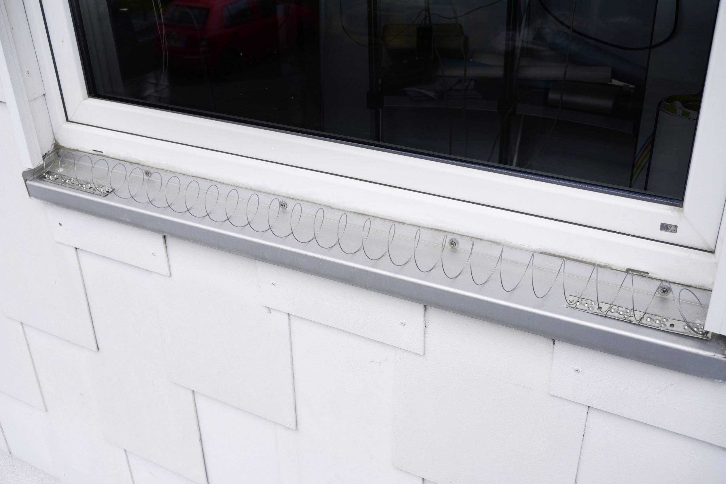 Fenster mit einem Anti-Tauben-Drahtsystem auf der Fensterbank, um Vögel fernzuhalten. Im Hintergrund sind Autos zu sehen.