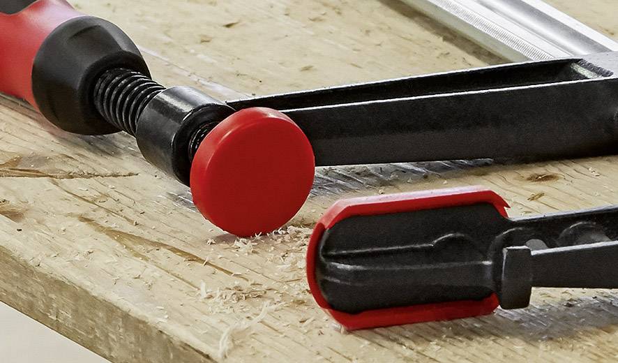 Eine Schraubzwinge liegt auf einer Holzwand, die von Holzspänen bedeckt ist. Die rote Grifffläche und der Spannmechanismus sind sichtbar.