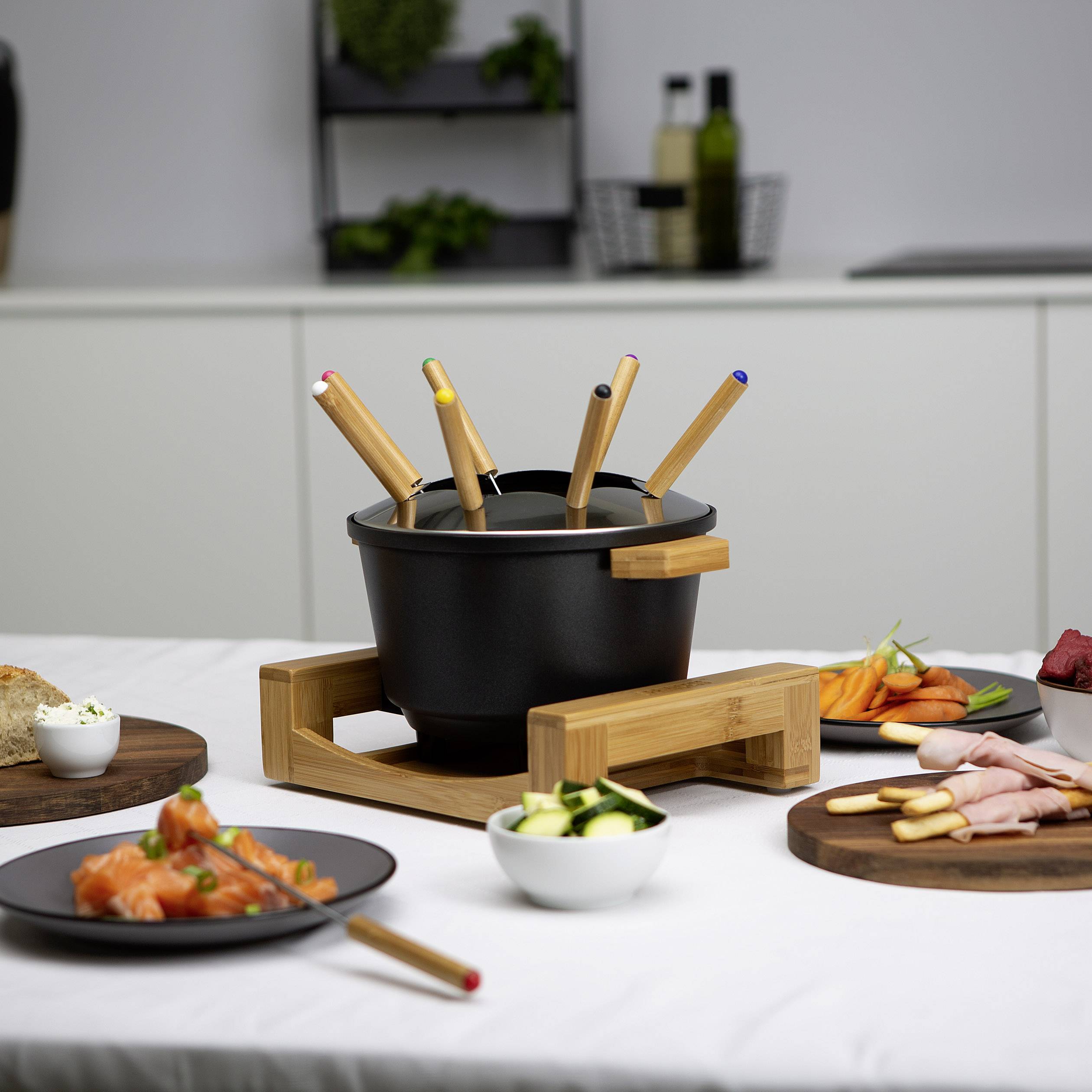Topf mit Käsefondue, umgeben von Tellern mit Lachs, Gemüse und Brot auf einem Tisch in moderner Küche.