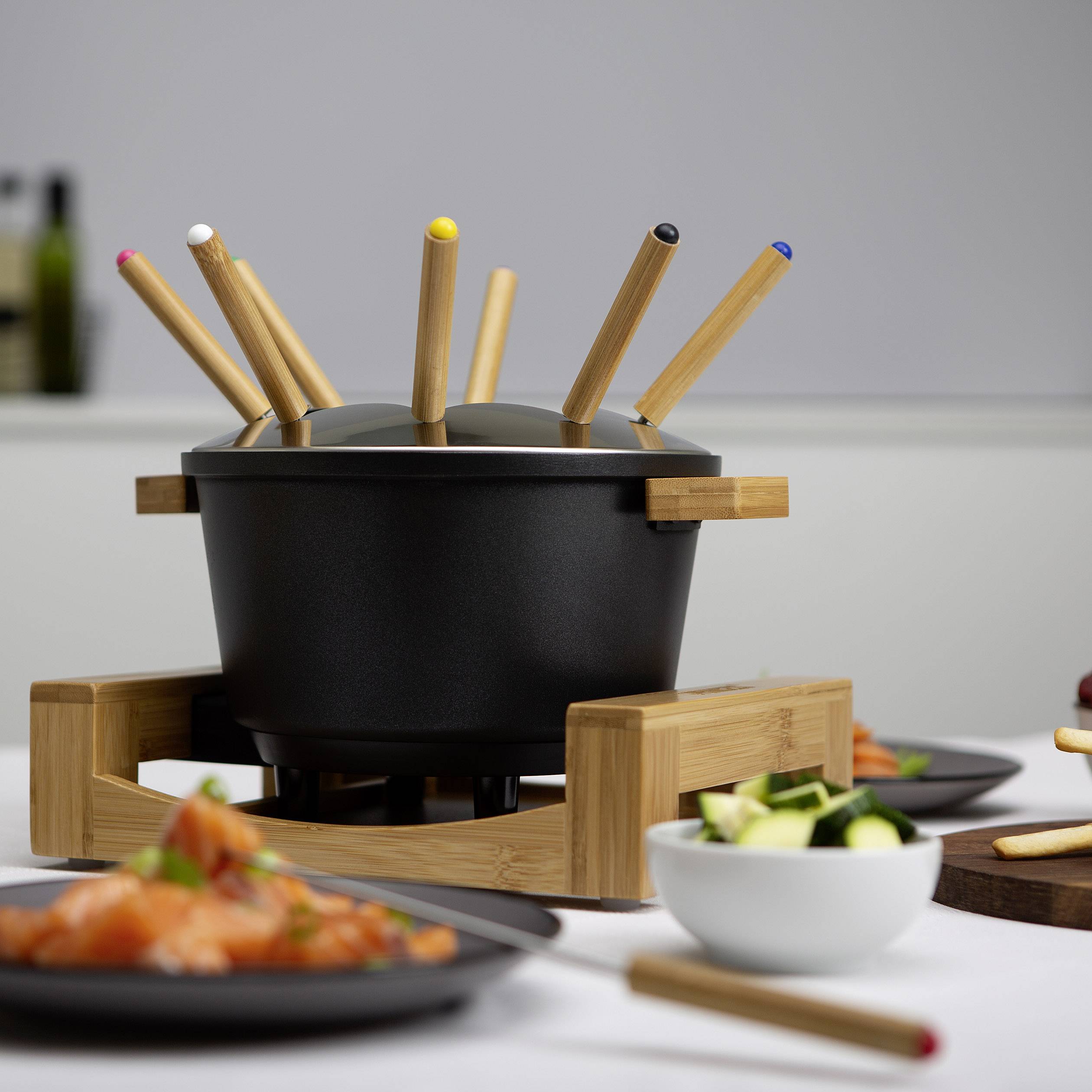 Schwarzer Fondue-Topf mit bunten Gabeln, umgeben von Tellern mit Gemüse und Lachs auf einem gedeckten Esstisch.