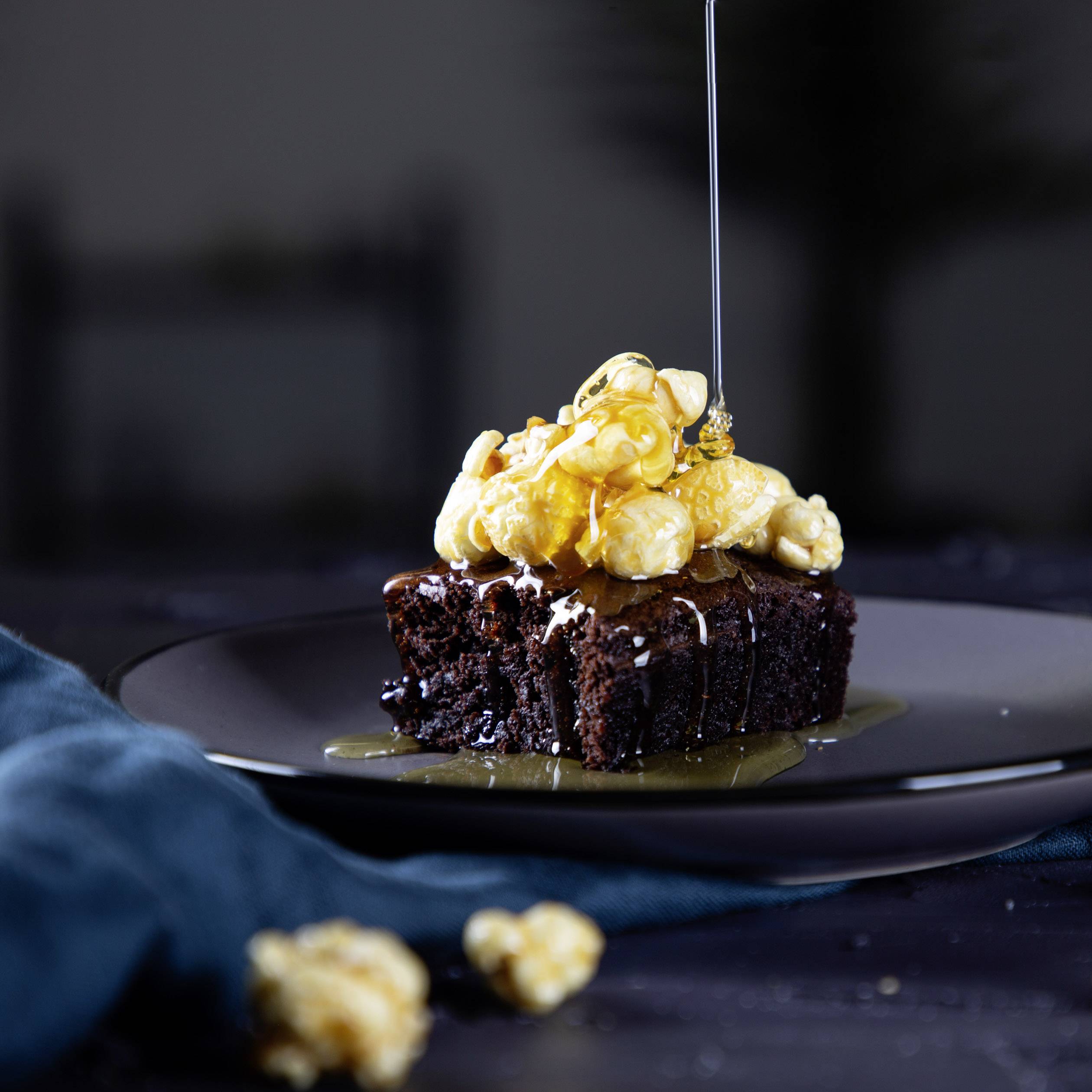 Ein Schokoladenkuchenstück mit Karamellpopcorn und Tropfen aus flüssigem Karamell, auf einem dunklen Teller angerichtet.