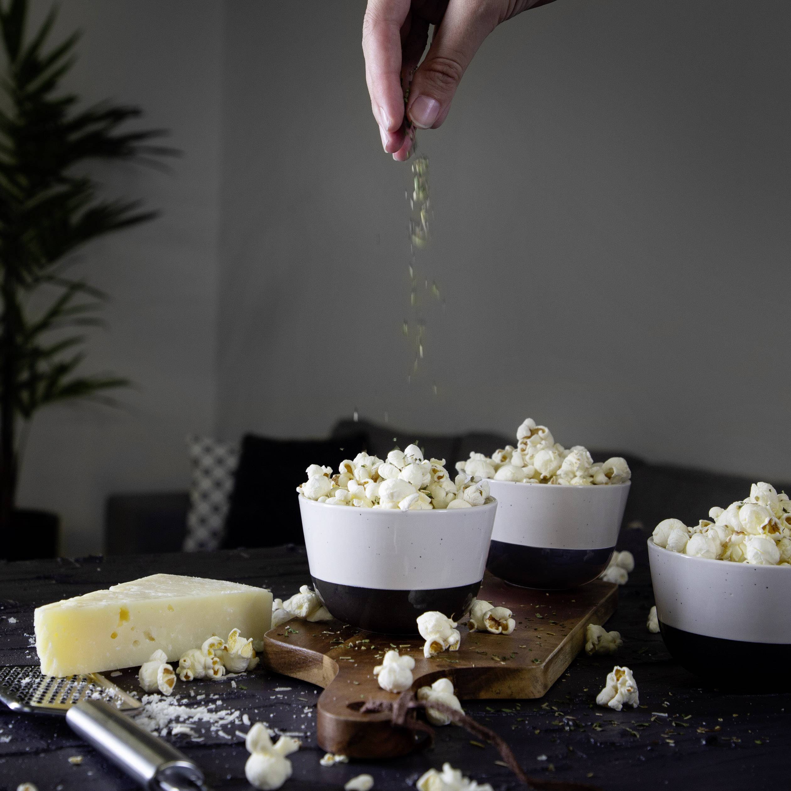 Eine Hand streut Kräuter über Popcorn in weißen Schalen auf einem Tisch mit Käse und einer Pflanze im Hintergrund.