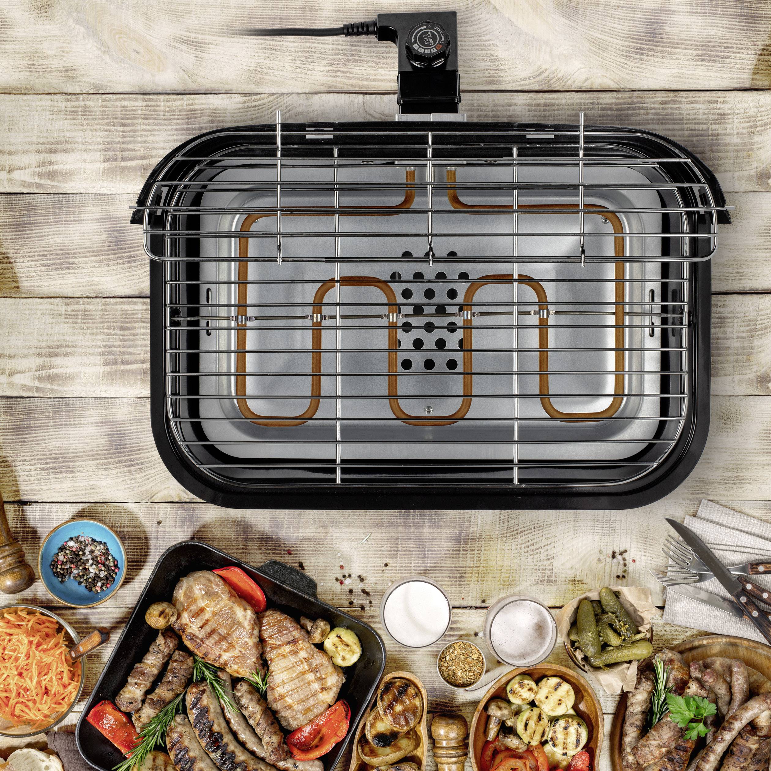 Elektrogrill mit Heizelement von oben, umgeben von gegrilltem Fleisch, Gemüse und Gewürzen auf einem Holztisch.