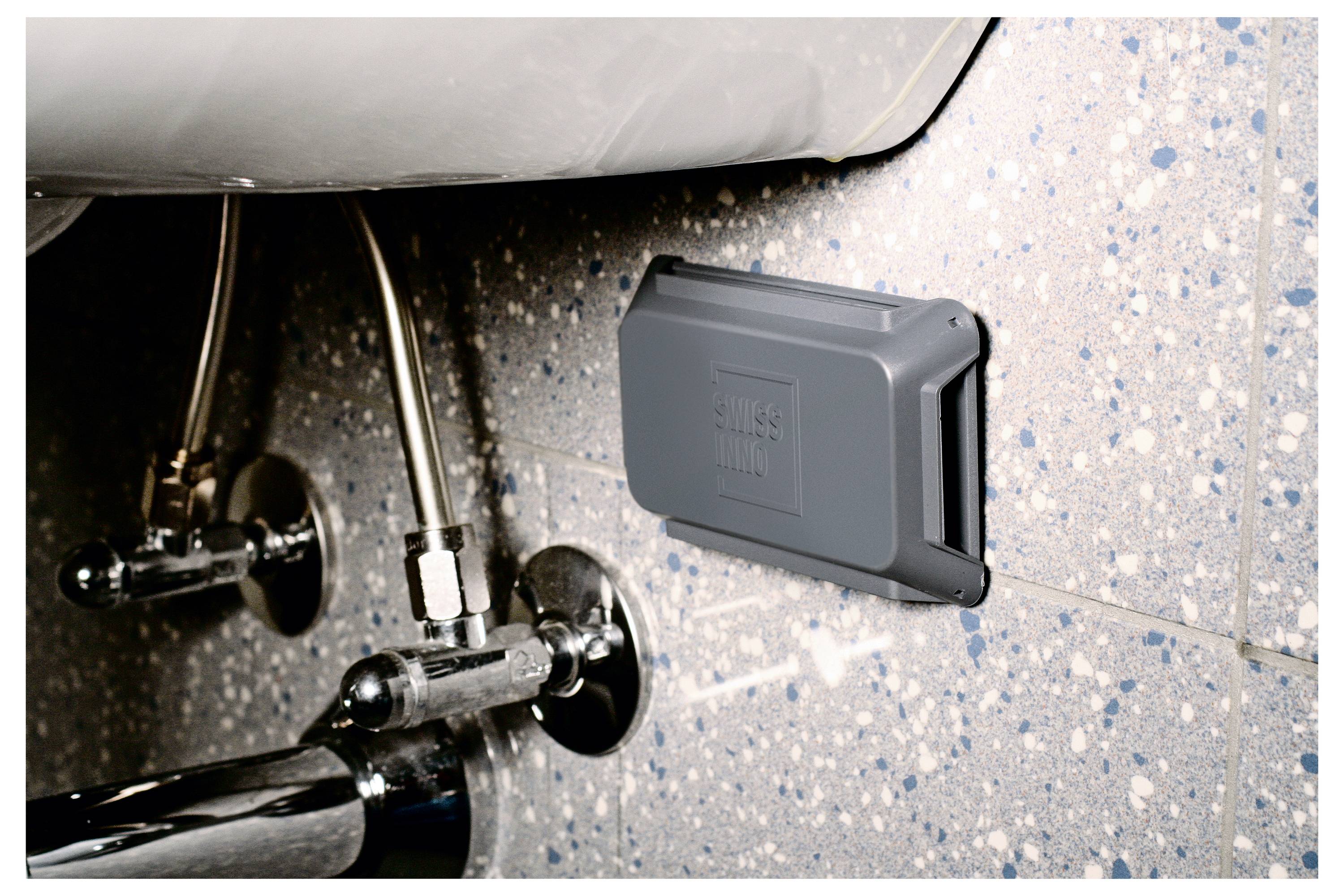 Ein grauer Kunststoffapparat, der an einer gefleckten Badezimmerwand in der Nähe von Klempnerinstallationen unter einem Waschbecken montiert ist.