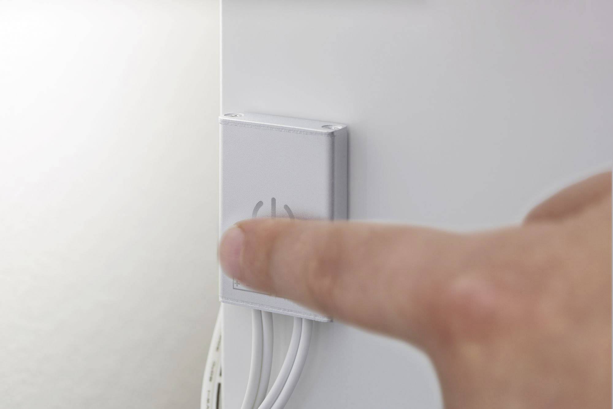 Ein Finger drückt einen großen, silbernen Knopf an der Seite eines weißen Geräts. Kabel sind unterhalb des Knopfes sichtbar.