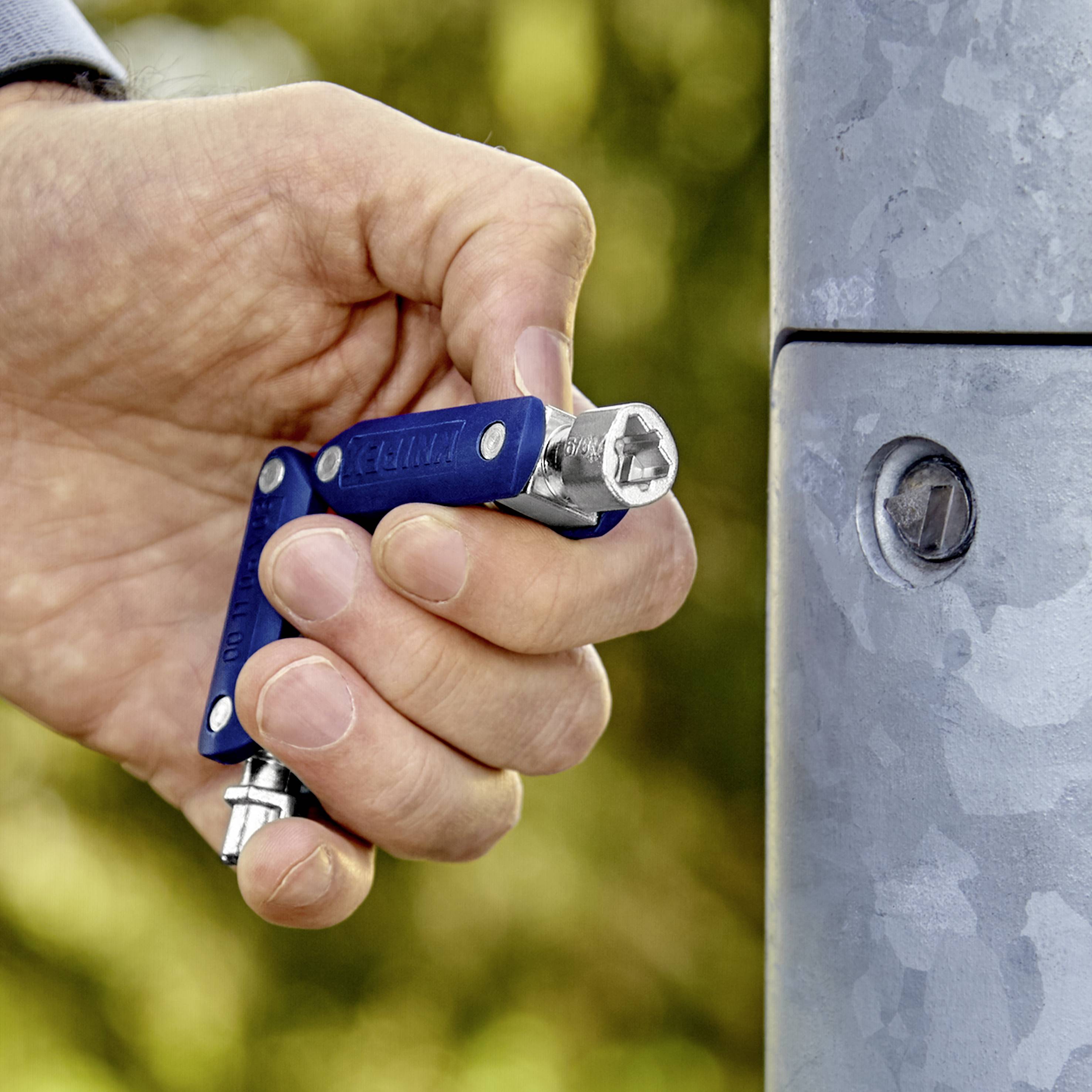 Eine Hand hält ein blaues Multi-Tool und zieht eine Schraube an einem Metallpfosten fest. Der Hintergrund ist unscharf und zeigt grüne Vegetation.
