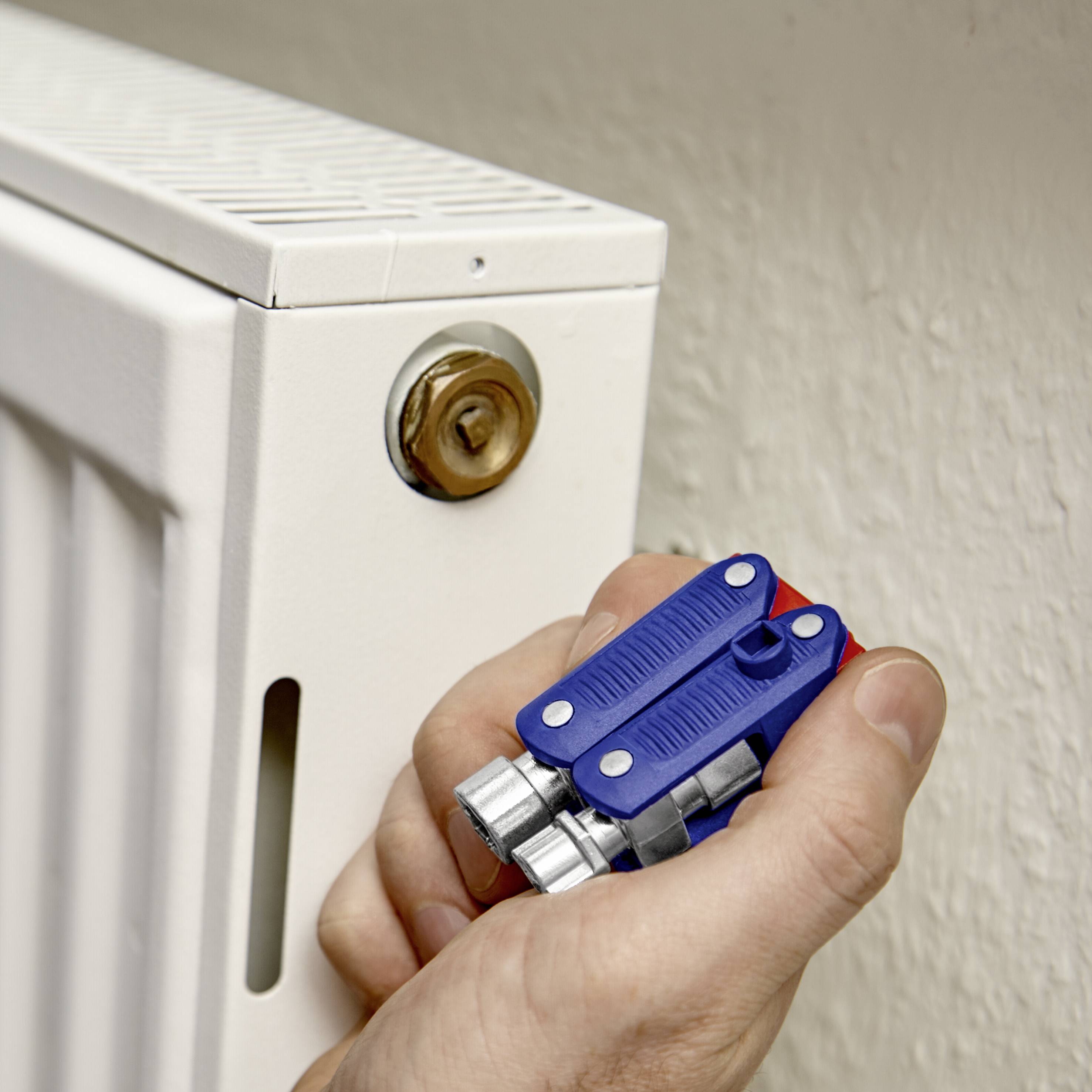 Eine Hand hält einen Heizkörperschlüssel vor einem weißen Heizkörper. Der Schlüssel wird am Ventil angesetzt, um Luft abzulassen.