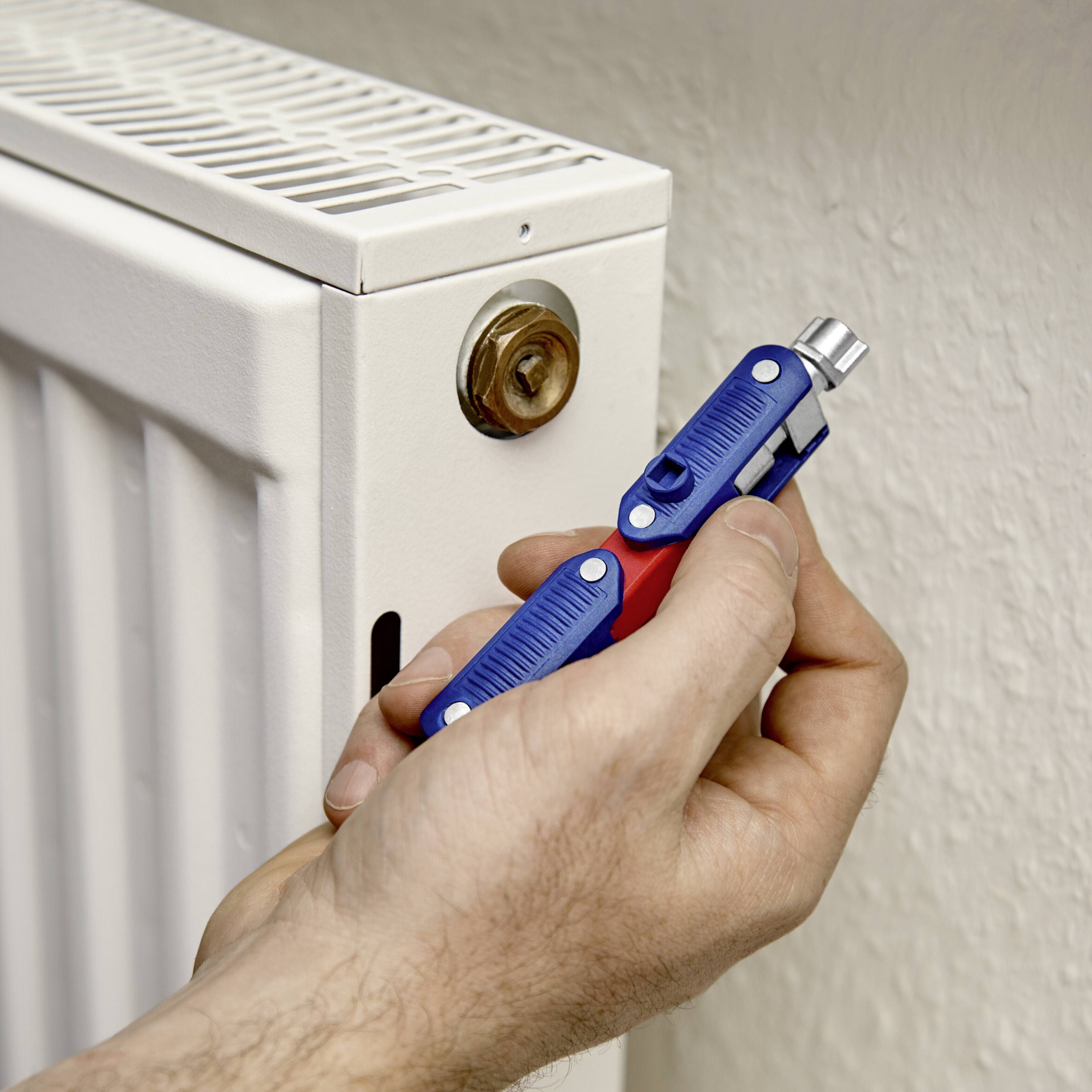 Eine Hand entlüftet einen Heizkörper mit einem Ventilschlüssel. Der Heizkörper ist an der Wand montiert und weiß lackiert.