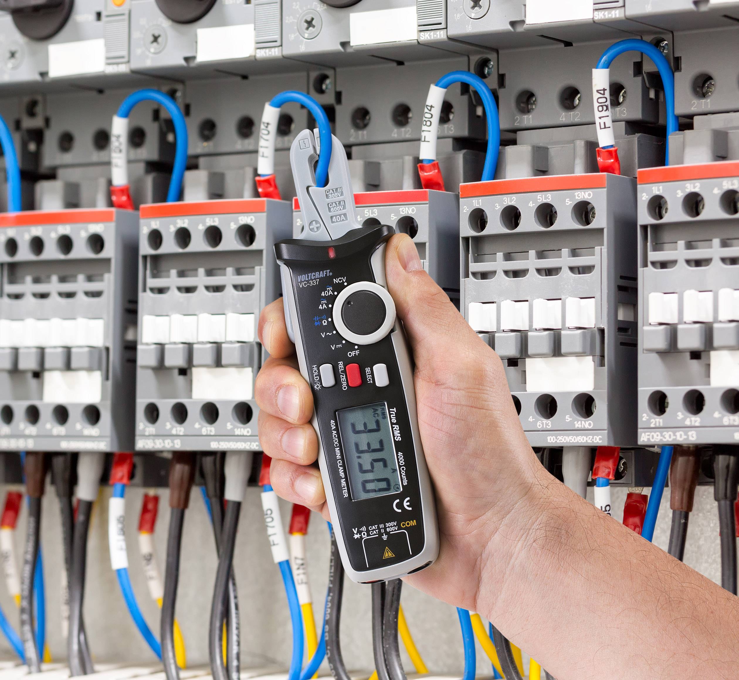 Eine Hand hält ein digitales Spannungsmessgerät vor einer geöffneten Schalttafel. Bunte Kabel sind mit verschiedenen Schaltern verbunden.