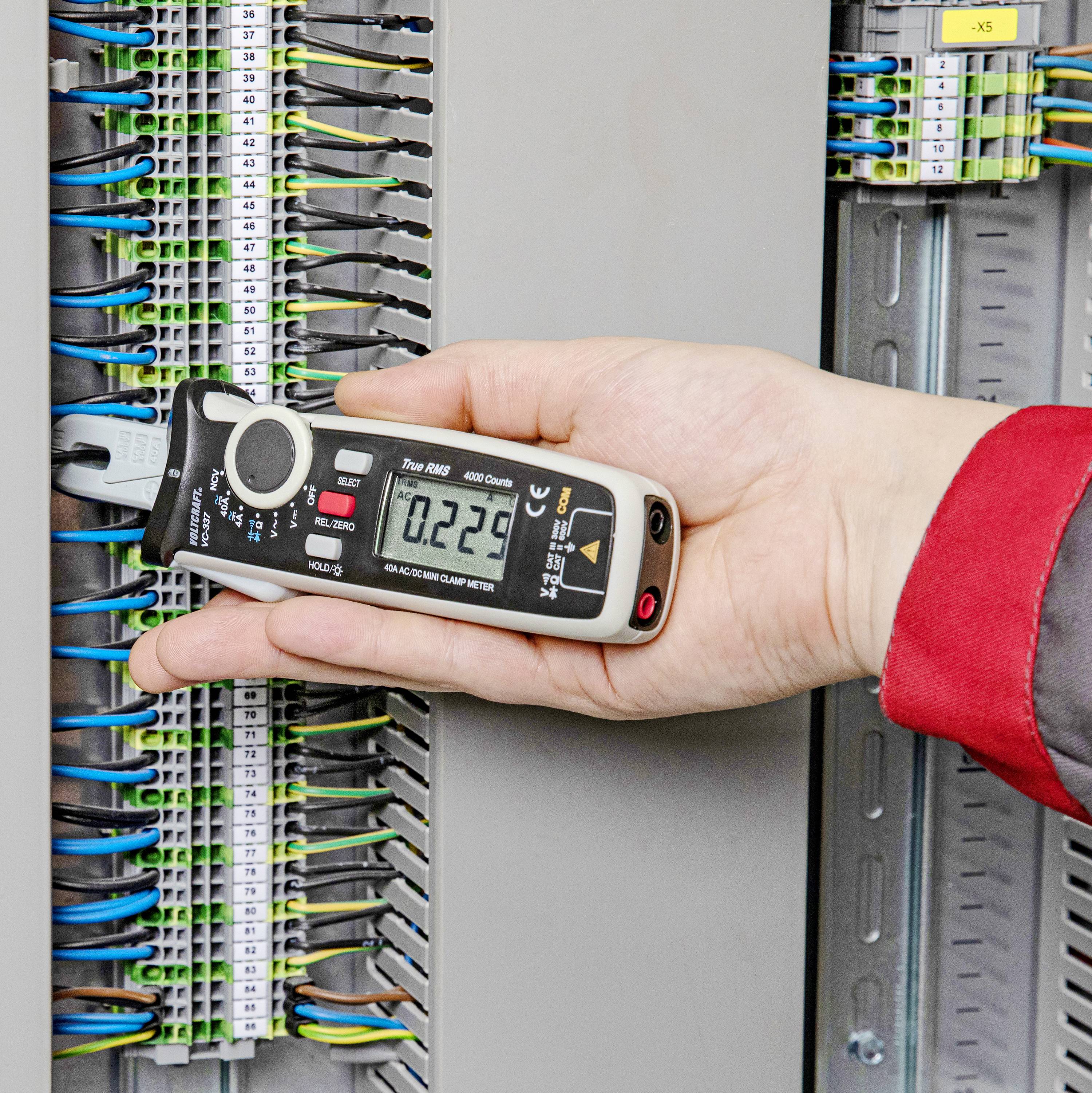 Eine Person hält ein digitales Messgerät an einem offenen Stromverteiler, um elektrische Signale zu messen. Kabel sind sichtbar angeschlossen.