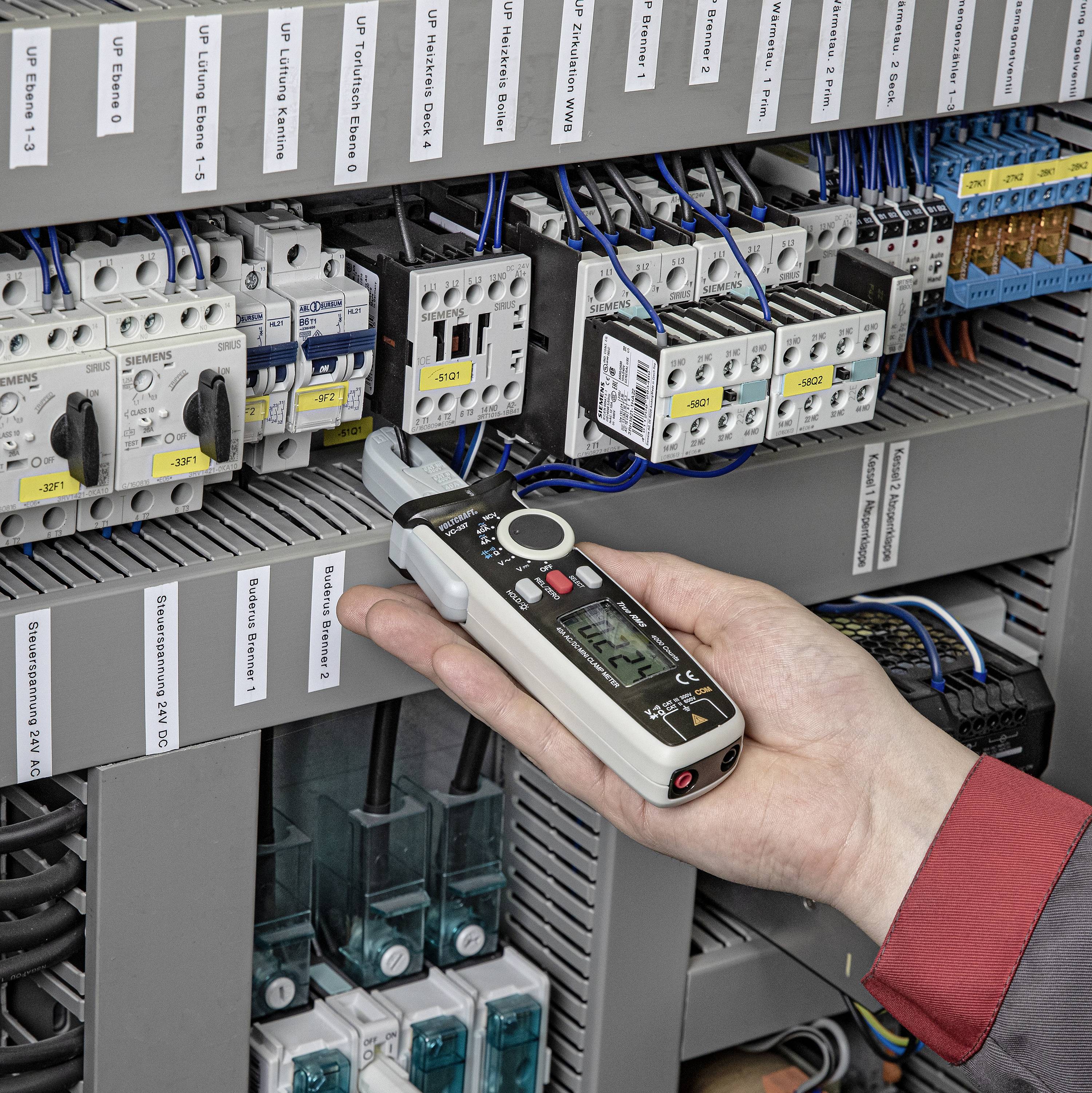 Eine Hand hält ein digitales Multimeter und misst den Strom in einem geöffneten Schaltschrank mit mehreren Verdrahtungen und Geräten.