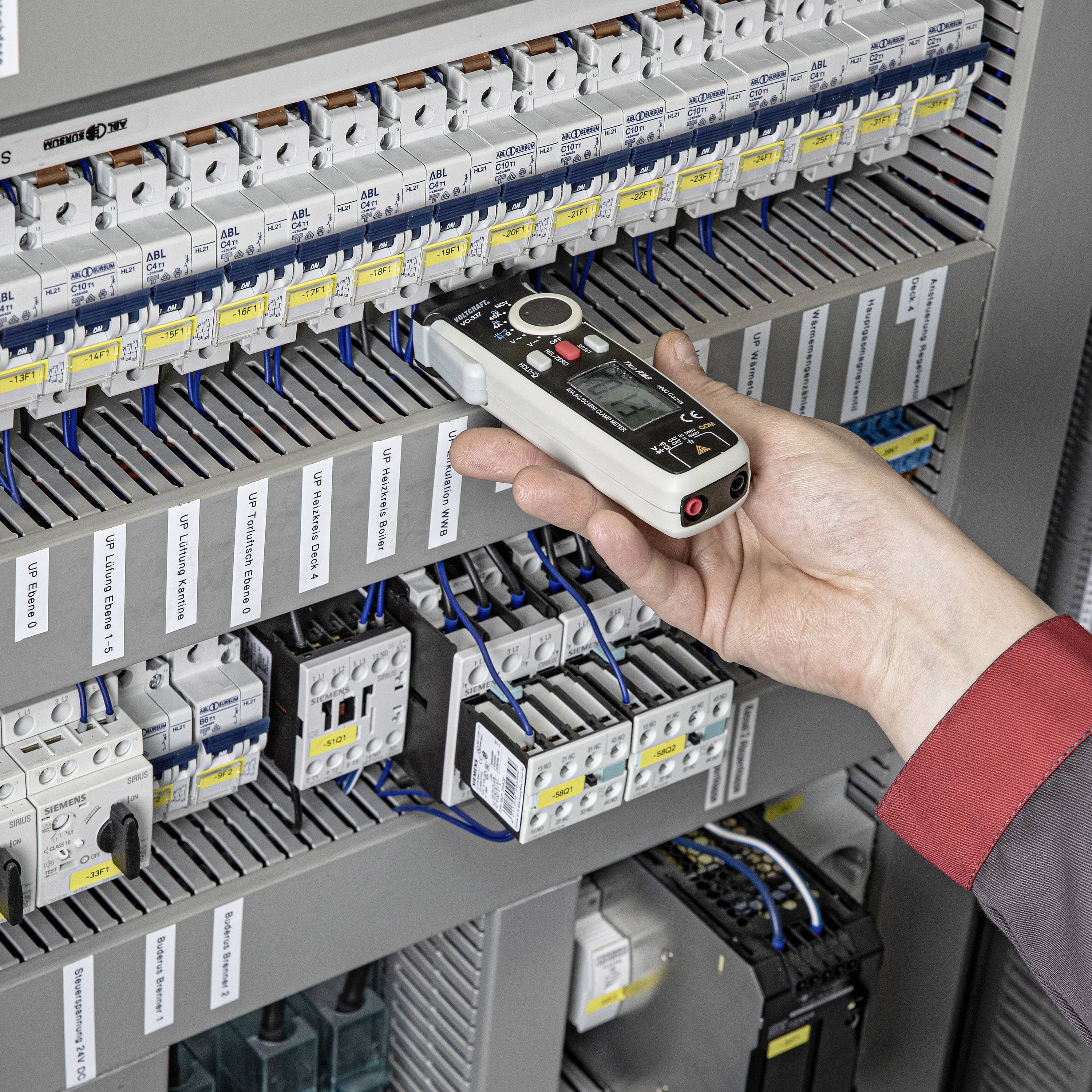 Eine Hand hält ein Messgerät vor einem Schaltschrank mit verschiedenen elektrischen Komponenten und Sicherungen zur Wartung oder Prüfung.