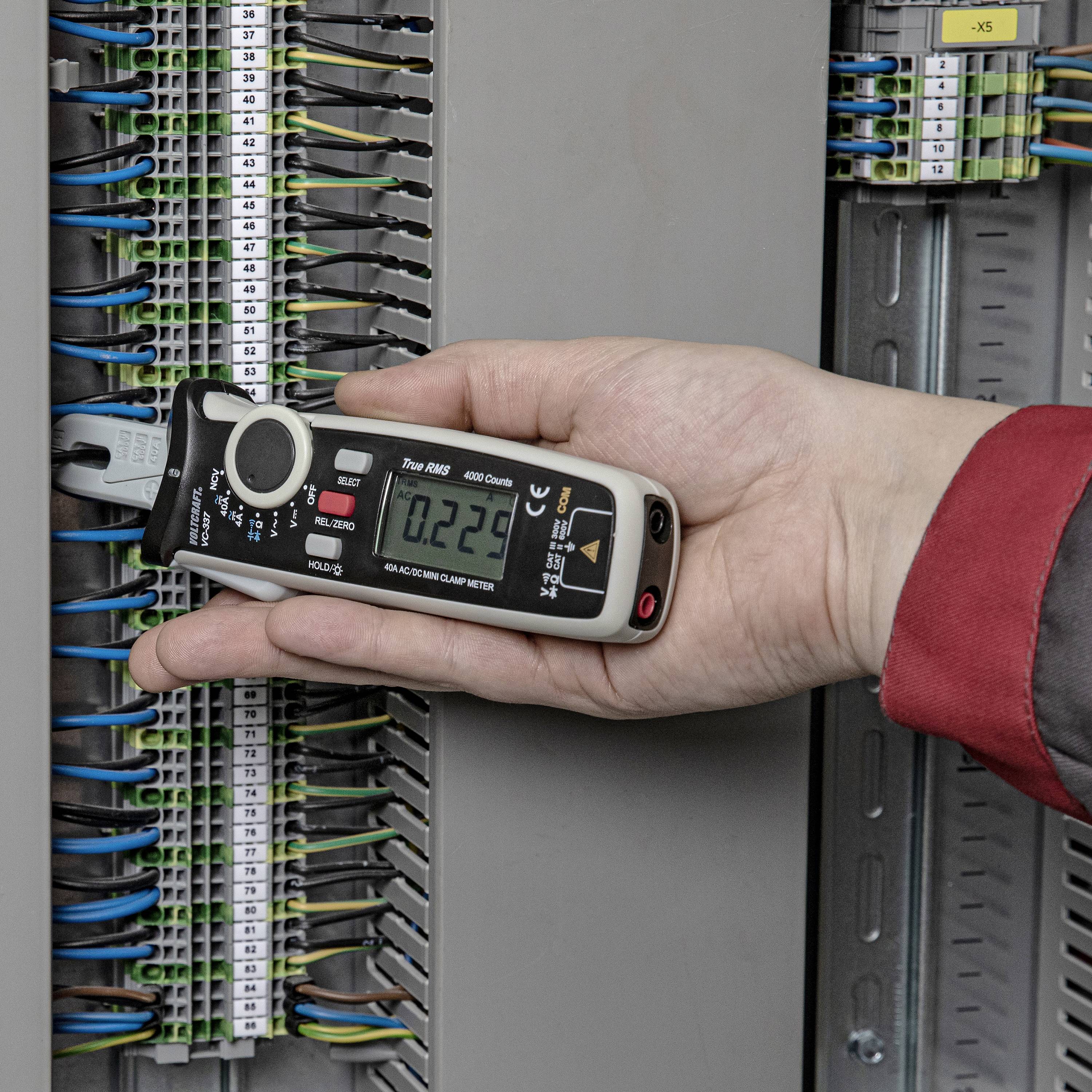 Eine Hand hält ein Multimeter, das an einem offenen Schaltschrank angeschlossen ist, um die elektrische Spannung zu messen.