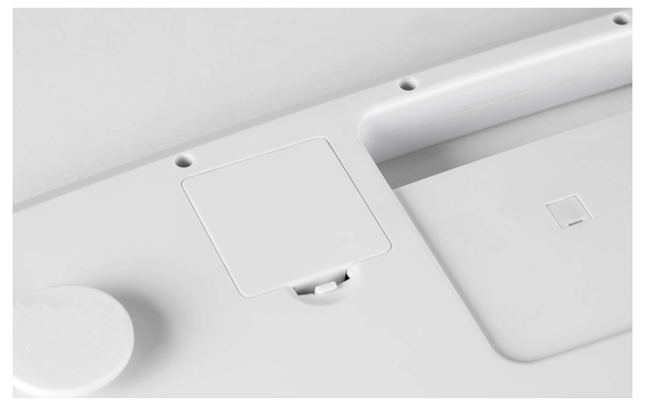 Weiße Unterseite eines elektronischen Geräts mit Batterieabdeckung und Aussparungen für Schrauben.