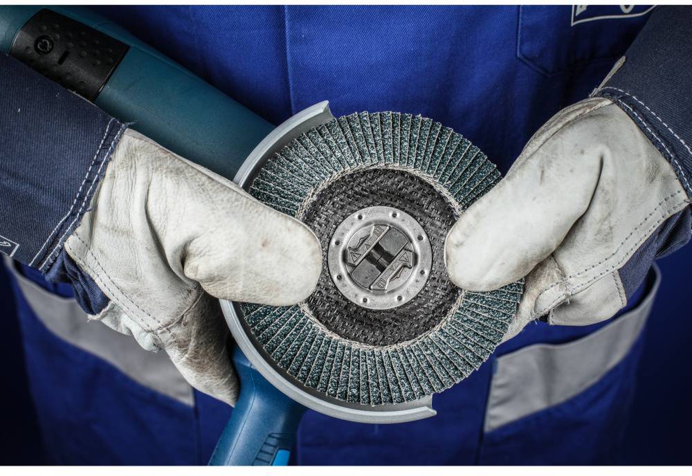 Ein Arbeiter in blauer Schutzkleidung und weißen Handschuhen hält eine elektrische Scheiben-Schleifmaschine.