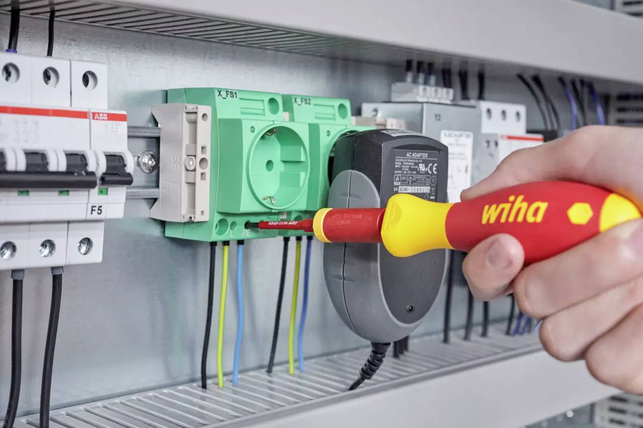 Eine Hand hält einen Wiha-Schraubendreher, während sie Drähte in einem Schaltschrank befestigt. Das Schaltfeld enthält verschiedene Schalter und Schaltungskomponenten.