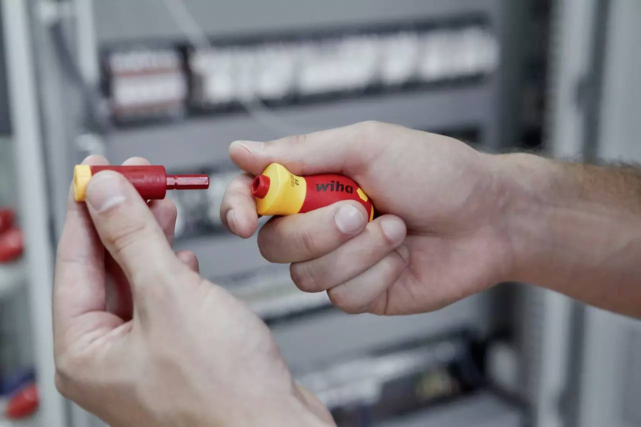 Zwei Hände halten ein rot und gelbes Werkzeug mit abnehmbarer Spitze, möglicherweise einen Schraubendreher, vor einem unscharfen Elektroschaltschrank.