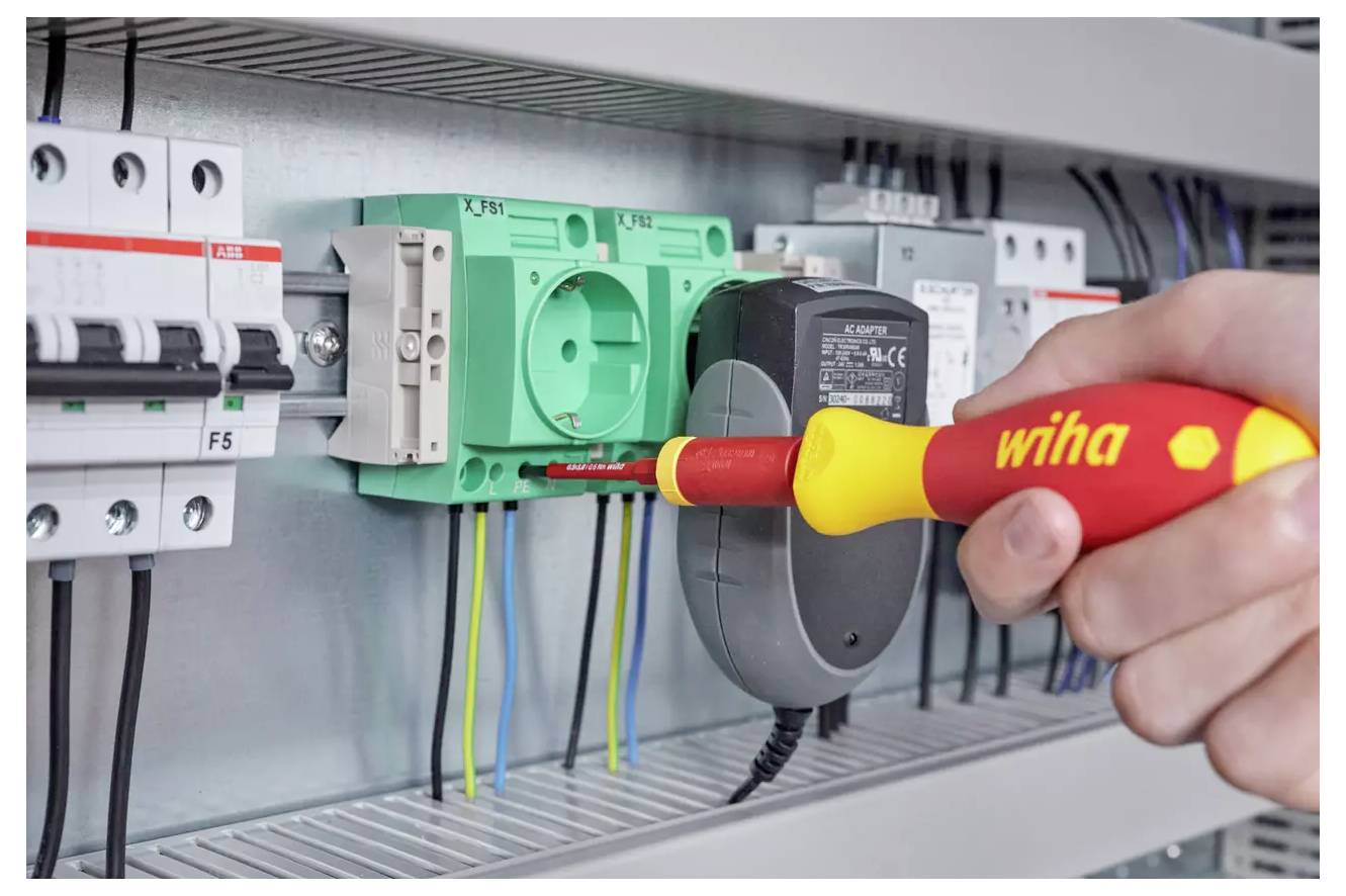 Eine Hand verwendet einen rot und gelb gefärbten Wiha-Schraubendreher, um ein grünes Elektrosteckdosenmodul innerhalb eines industriellen Steuerschalters festzuziehen.