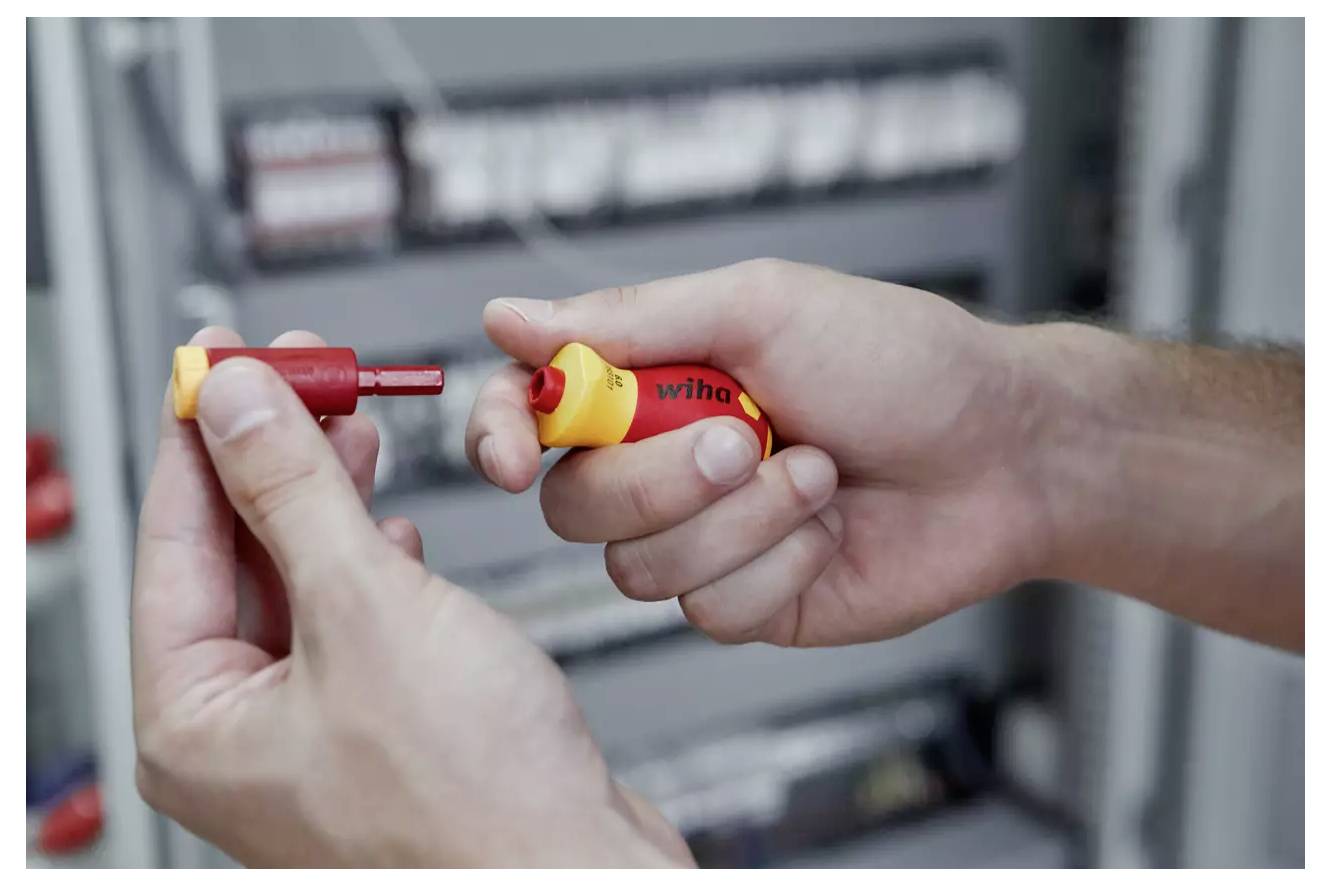 Eine Person hält einen rot-gelben Schraubendreher und montiert oder demontiert dessen Bits, während elektrische Ausrüstung im Hintergrund verschwommen ist.