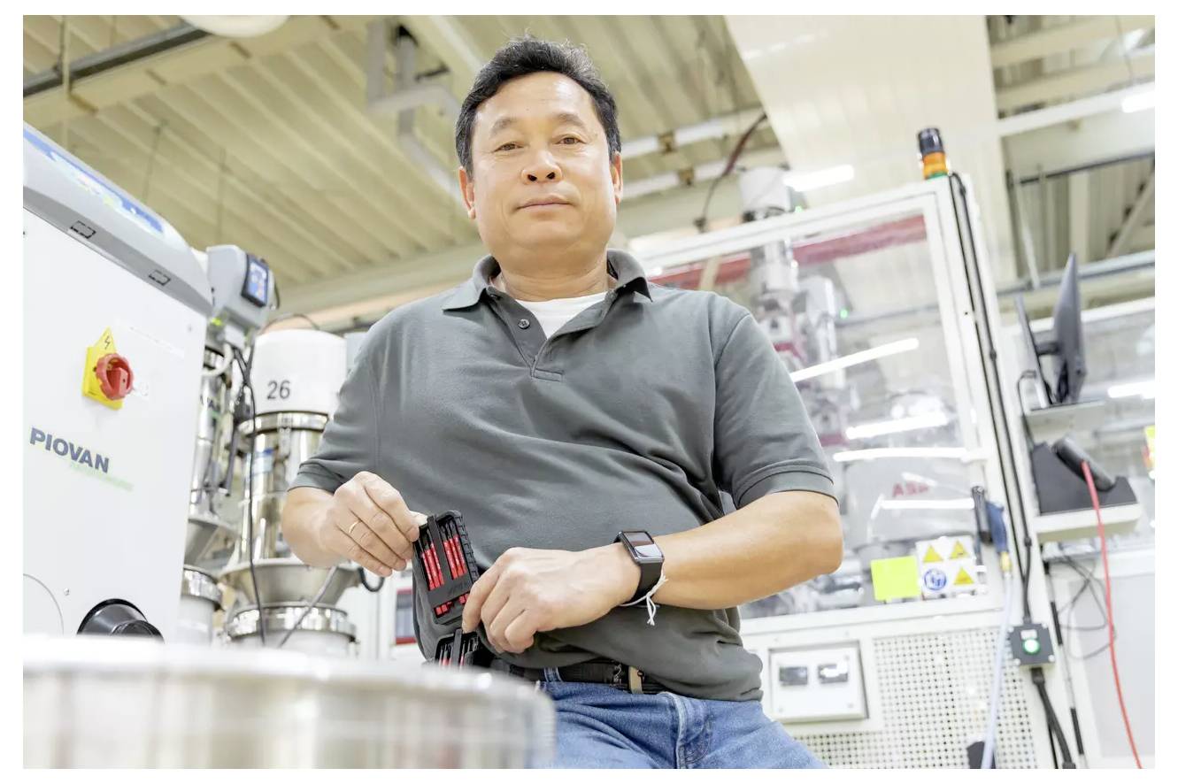 Eine Person in einer Fabrikumgebung hält kleine Ausrüstung, wirkt konzentriert. Maschinen und Bedienfelder umgeben sie, was auf eine Industrieumgebung hindeutet.