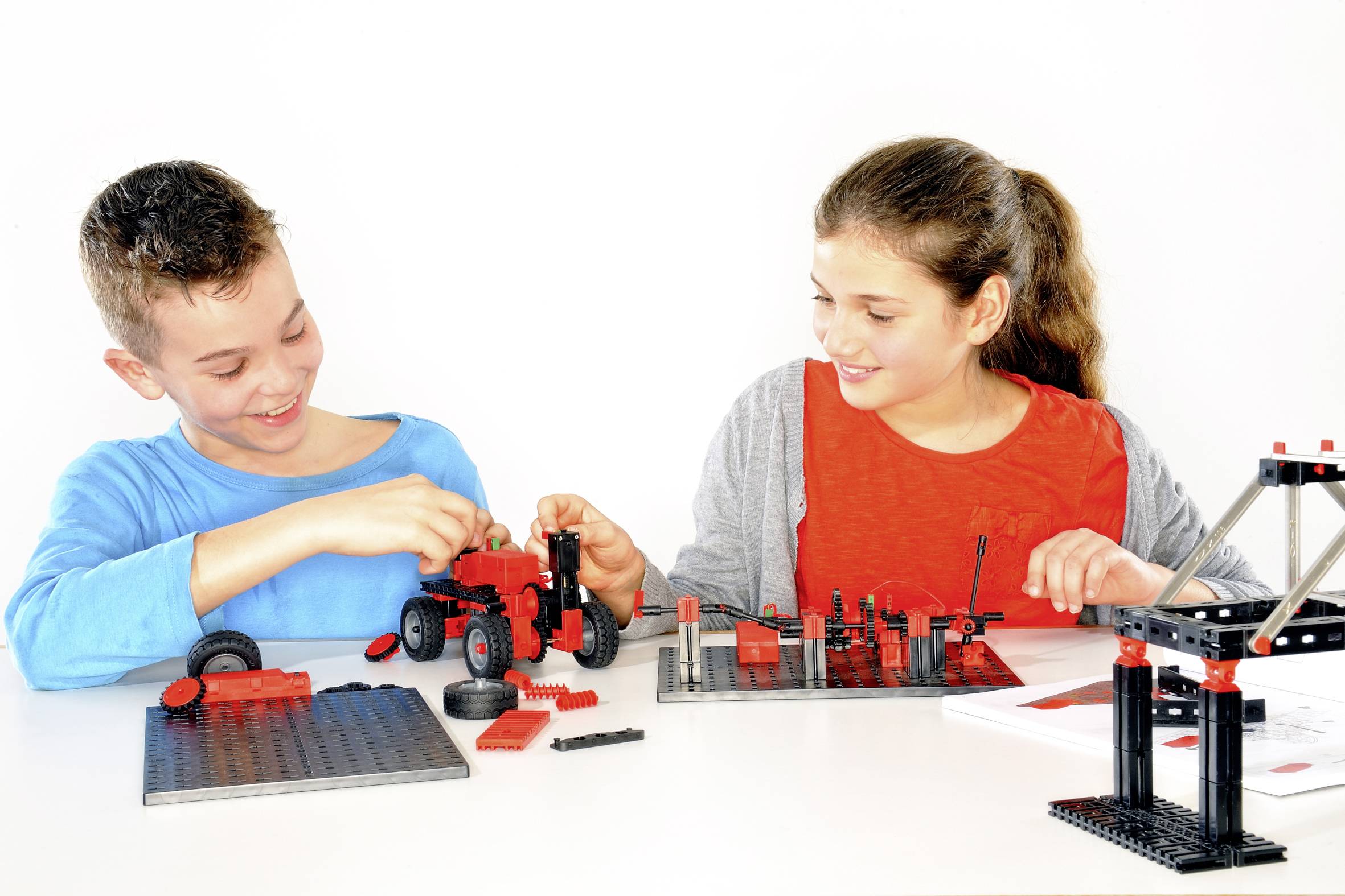 Zwei Kinder bauen lächelnd mit roten und schwarzen Bausteinen Modelle an einem Tisch. Sie sind in eine kreative Aktivität vertieft.