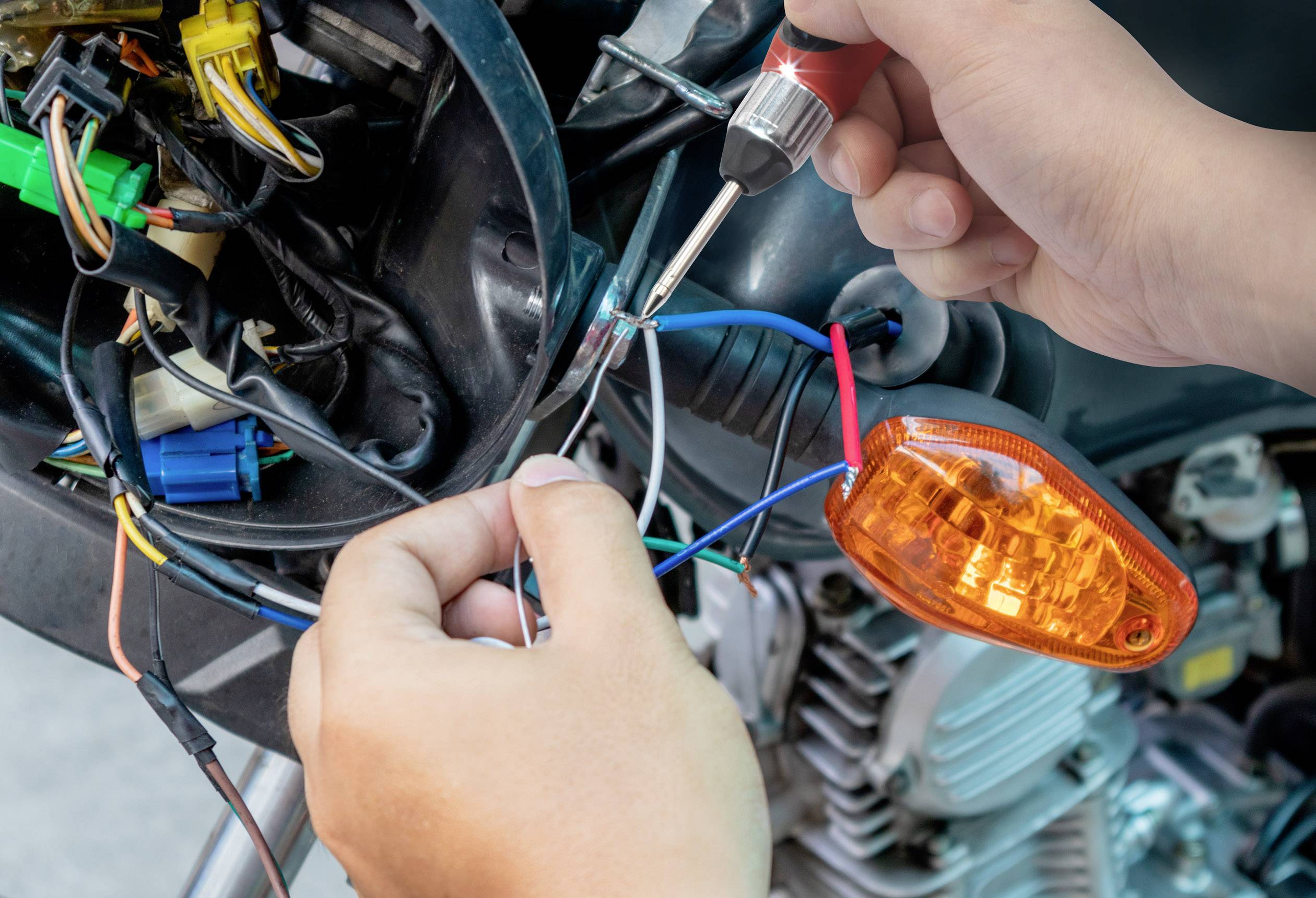 Eine Person nutzt einen Schraubendreher, um Drähte eines Motorrads zu verbinden. Ein orangefarbenes Blinklicht ist sichtbar.