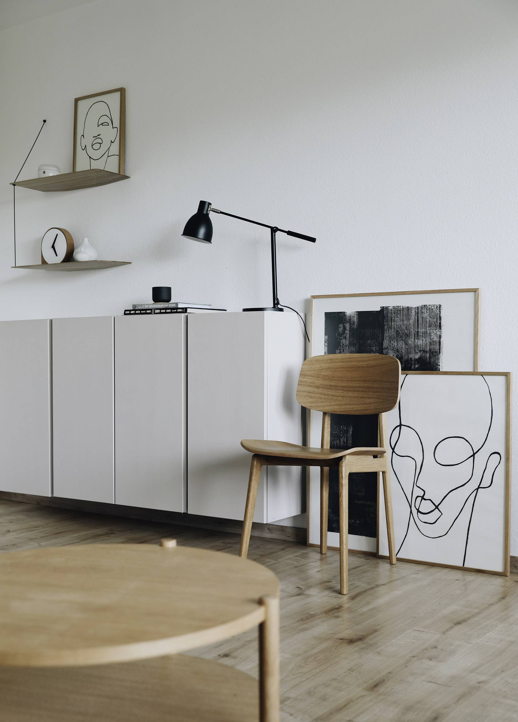 Ein minimalistisch eingerichtetes Zimmer mit Holzstuhl, hellen Regalen und moderner Kunst an der Wand. Eine Stehlampe steht auf dem Regal.