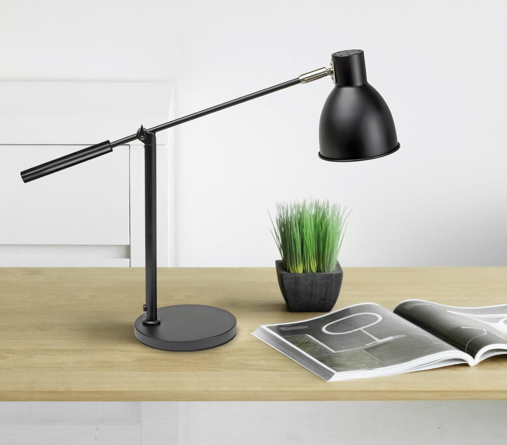 Schreibtisch mit schwarzer Schreibtischlampe, aufgeschlagenem Magazin und kleinem grünen Ziergras in einem Topf.