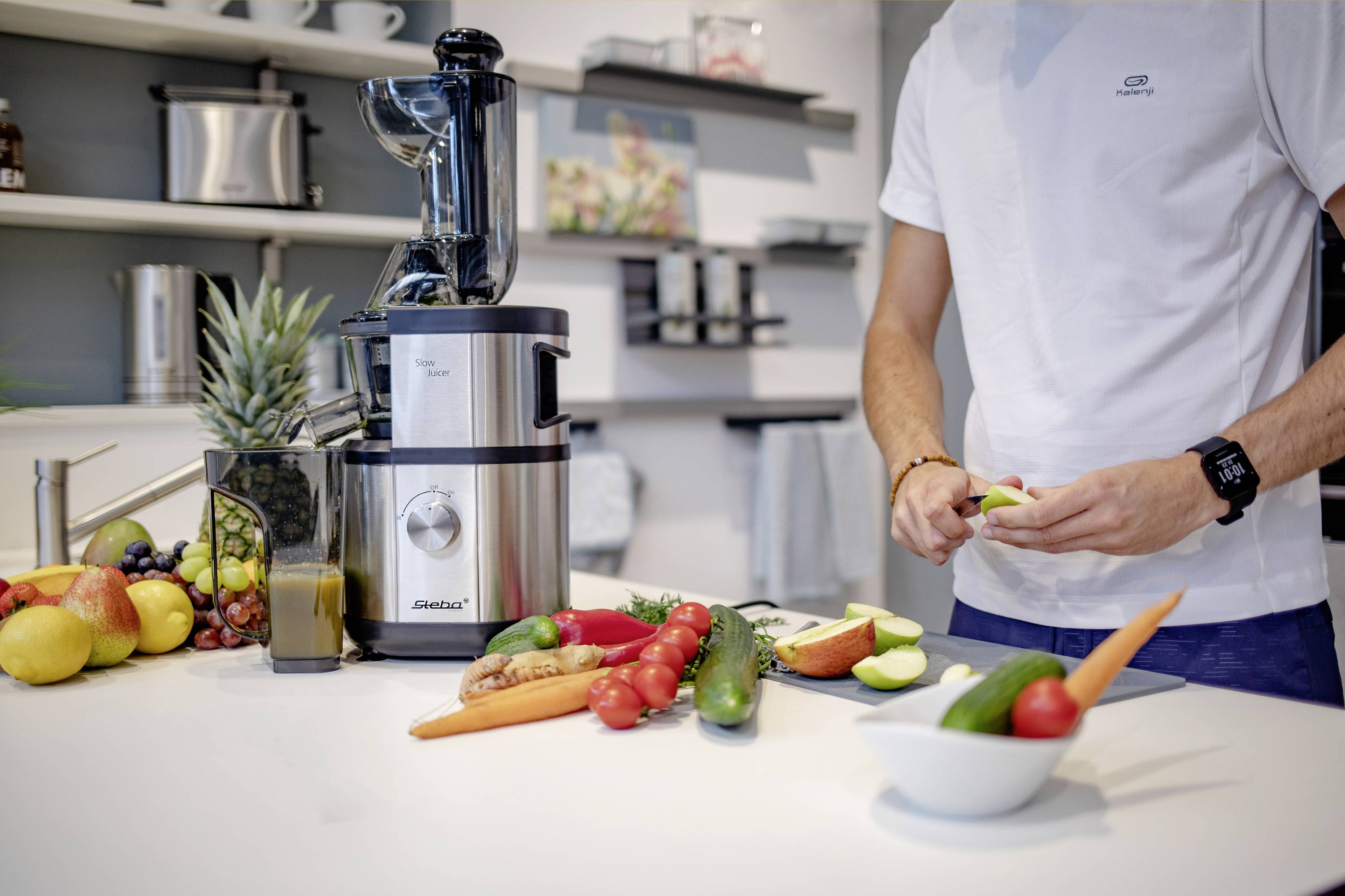 Eine Person bereitet in einer modernen Küche Obst und Gemüse neben einem Entsafter vor, umgeben von verschiedenen Früchten.