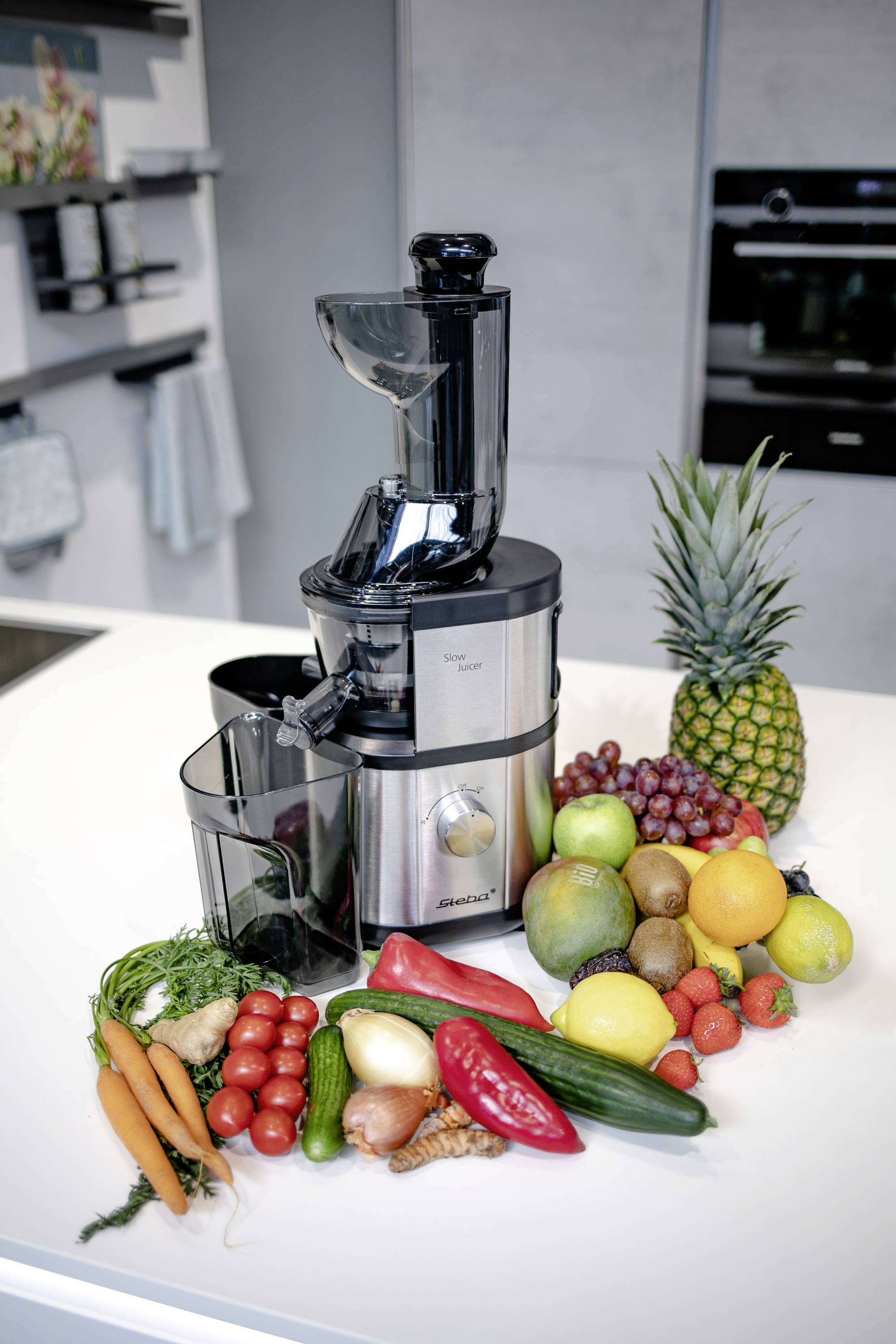 Entsafter auf einer Küchenarbeitsplatte, umgeben von frischem Obst und Gemüse wie Ananas, Trauben, Zitronen, Karotten und Paprika.