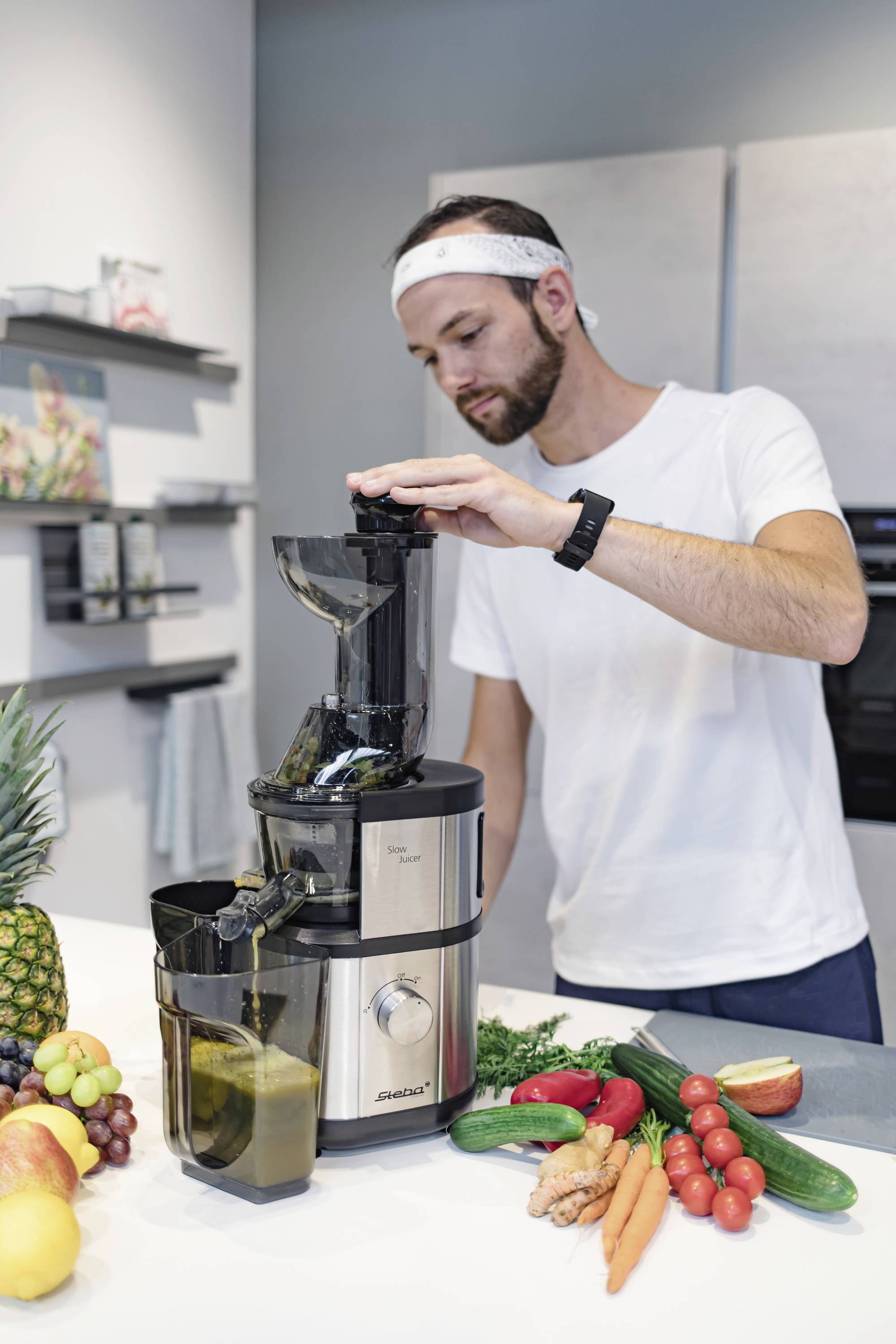 Ein Mann bereitet mit einem Entsafter Säfte zu. Auf der Küchenarbeitsplatte liegen Obst und Gemüse. Er trägt ein weißes T-Shirt und ein Stirnband.