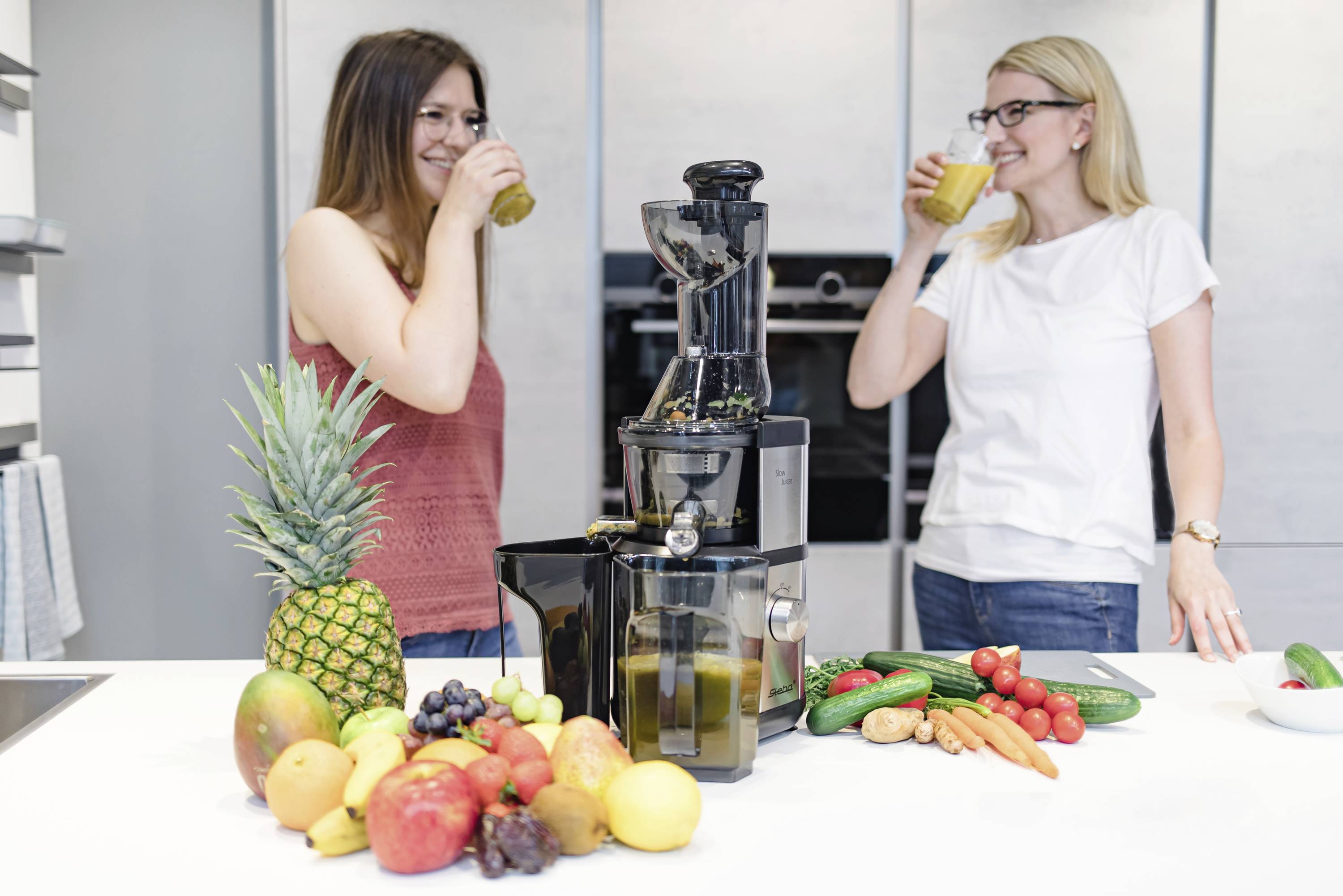 Zwei Frauen trinken Saft in einer modernen Küche. Auf der Theke steht ein Entsafter, umgeben von Obst und Gemüse wie Ananas und Tomaten.