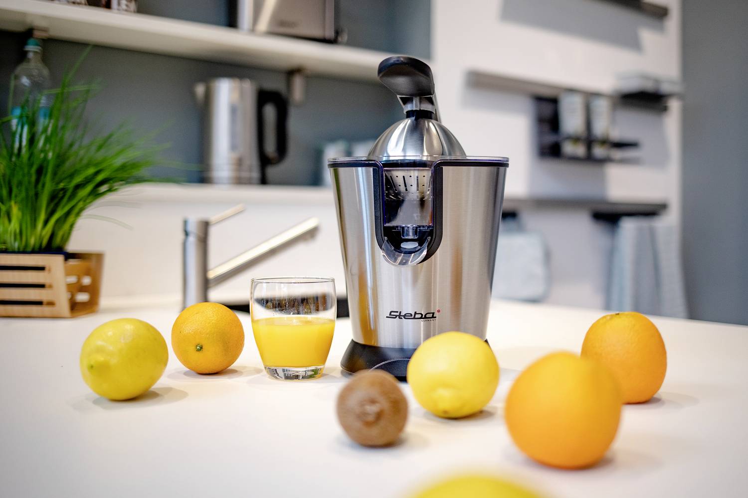 Eine Edelstahl-Zitruspresse auf einem weißen Küchentisch, umgeben von Orangen, Zitronen und einem Glas frischem Saft. Im Hintergrund Küchenutensilien.