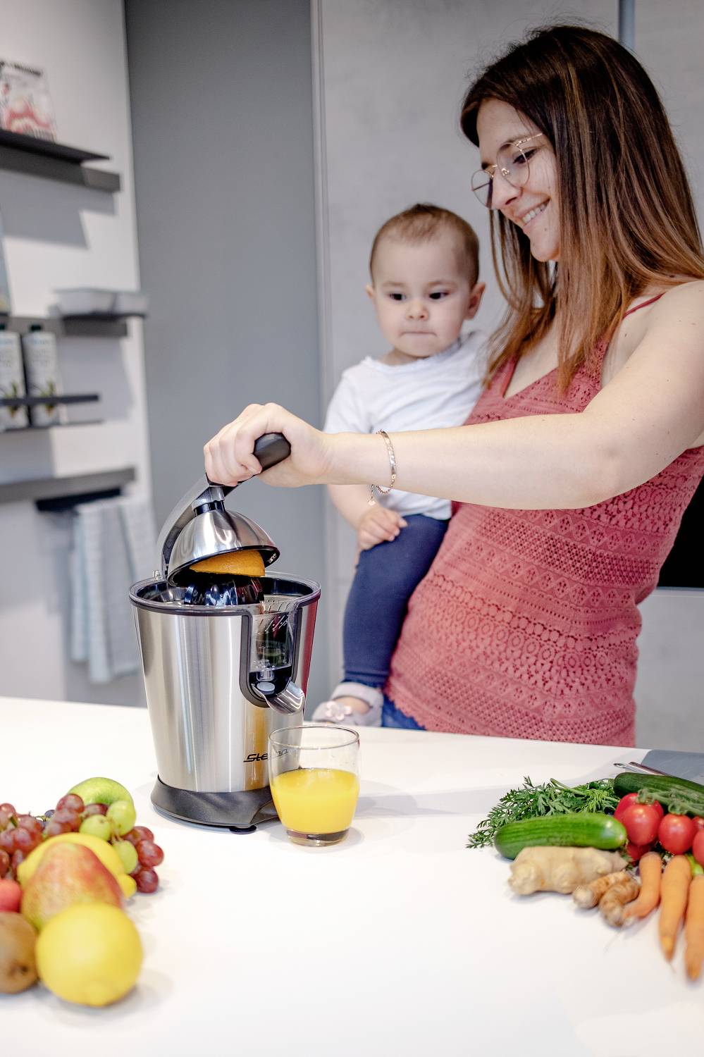 Eine Frau hält ein Baby im Arm und presst Orangen mit einem Entsafter aus. Auf dem Tisch liegen Früchte und Gemüse.