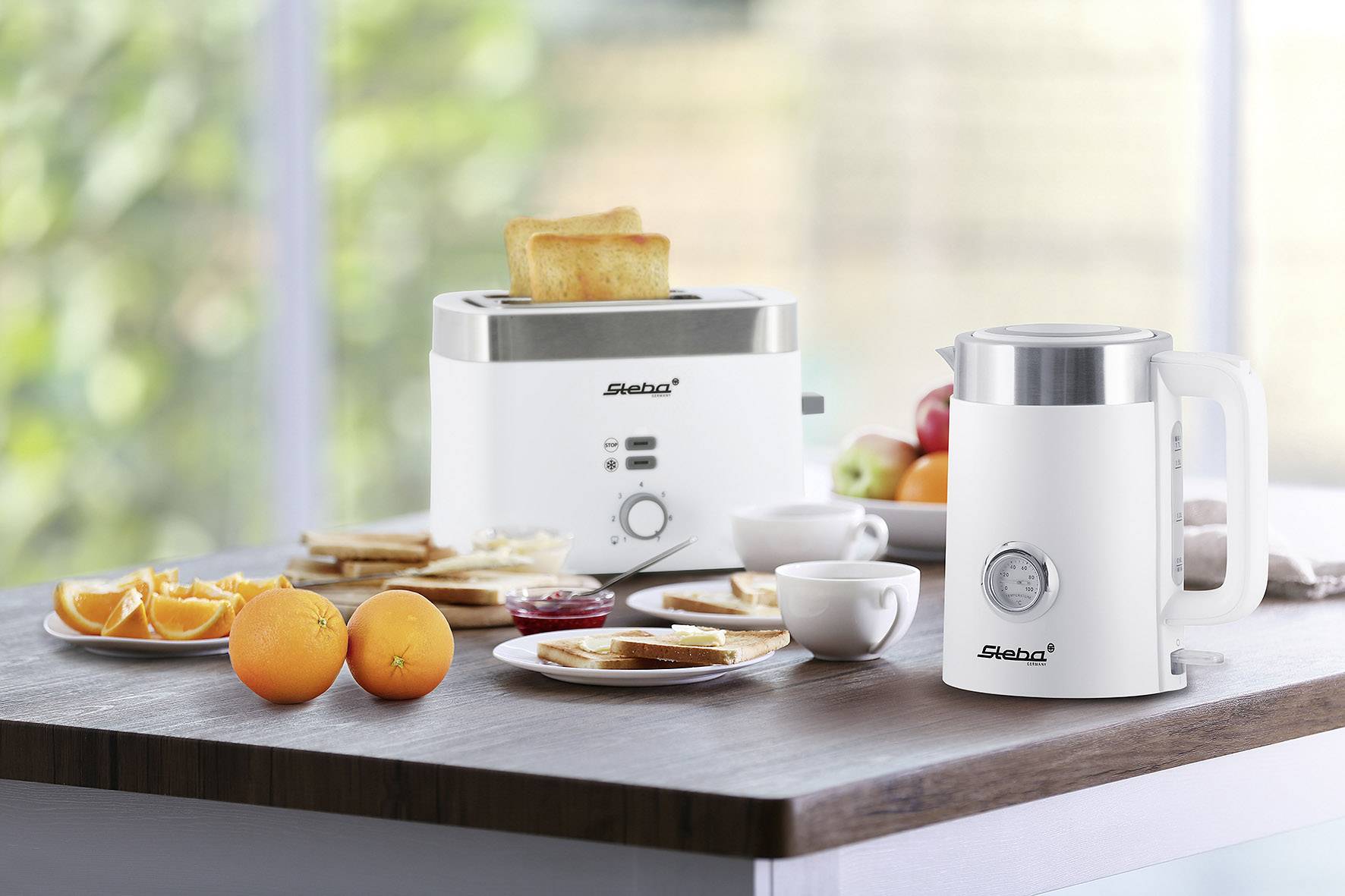 Ein Frühstückstisch mit einem Toaster, der Toastscheiben zubereitet, einer Teekanne, verschiedenen Früchten, Marmelade und Tassen.