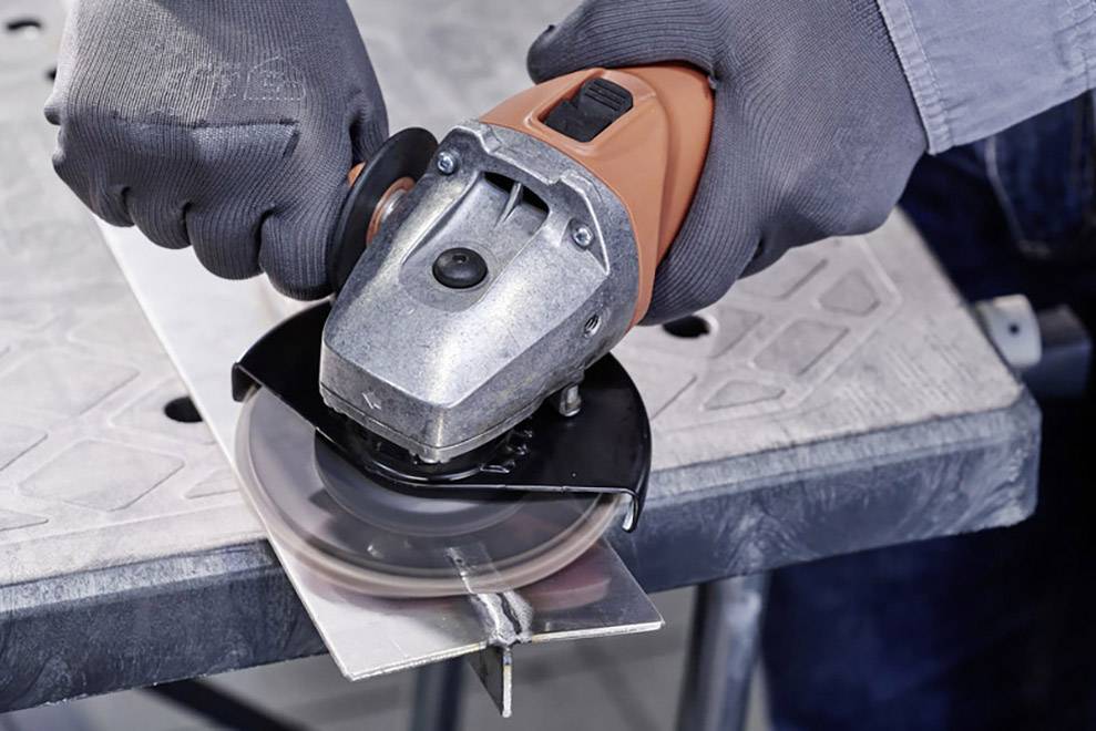 Person hält einen Winkelschleifer zum Schneiden eines Metallstücks auf einem Tisch. Arbeiter trägt Arbeitshandschuhe.