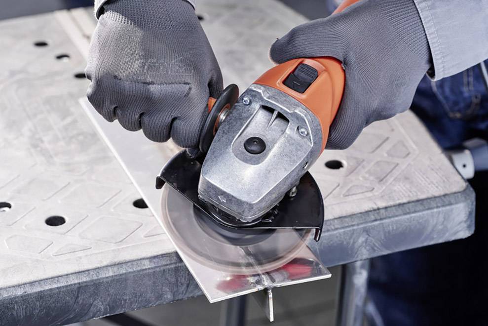 Person mit grauen Handschuhen schneidet ein Stück Metall mit einer orangefarbenen Winkelschleifmaschine auf einem Tisch.