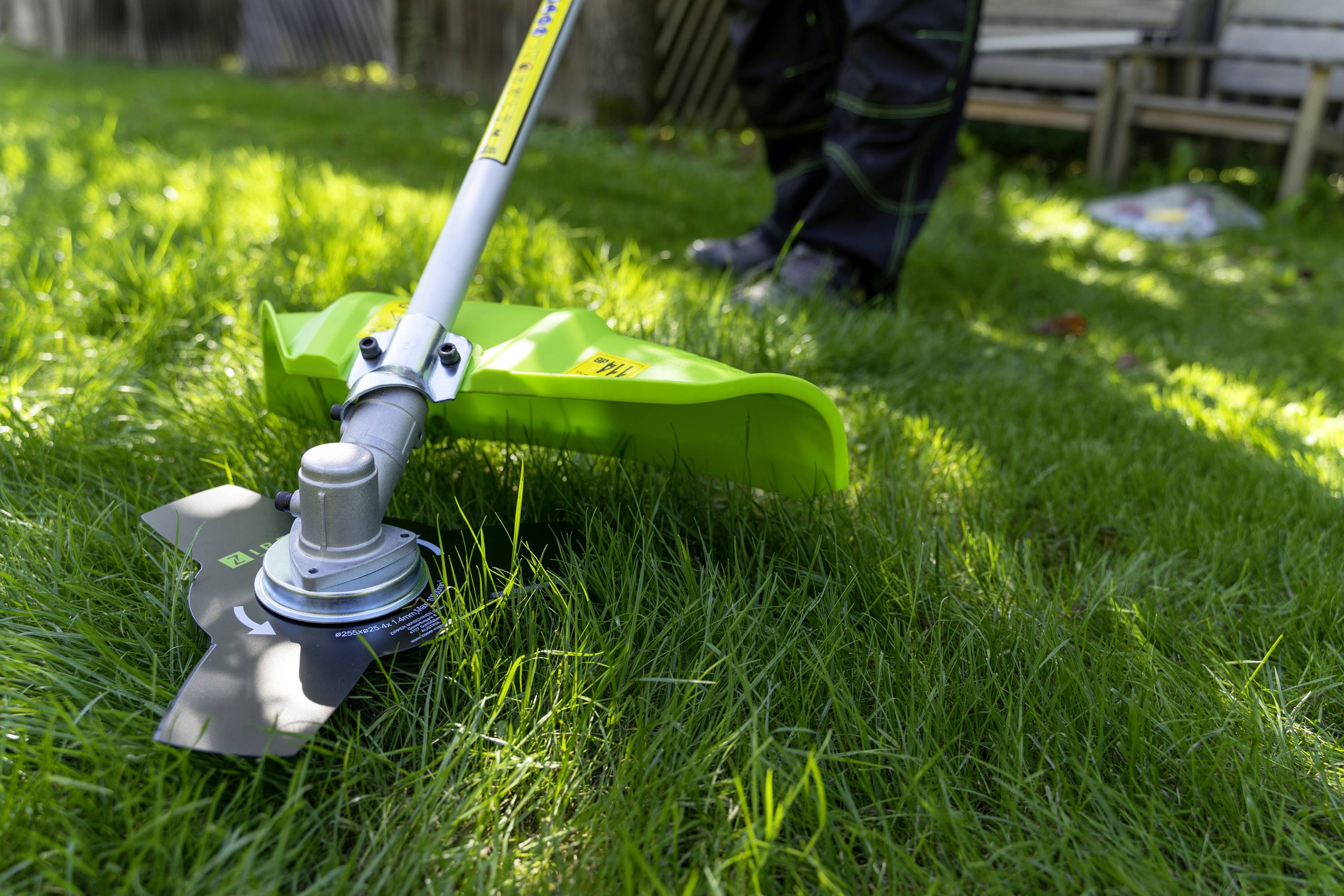 Ein Rasentrimmer schneidet Gras in einem Garten. Fokus liegt auf dem Schneidkopf, während im Hintergrund unscharf ein Holztisch zu sehen ist.