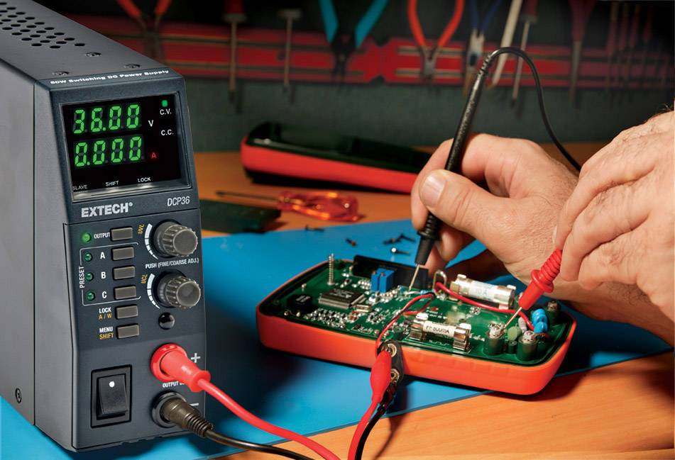 Nahaufnahme eines digitalen Multimeters und eines Oszilloskops auf einem Tisch. Eine Hand misst Strom an einer Leiterplatte.