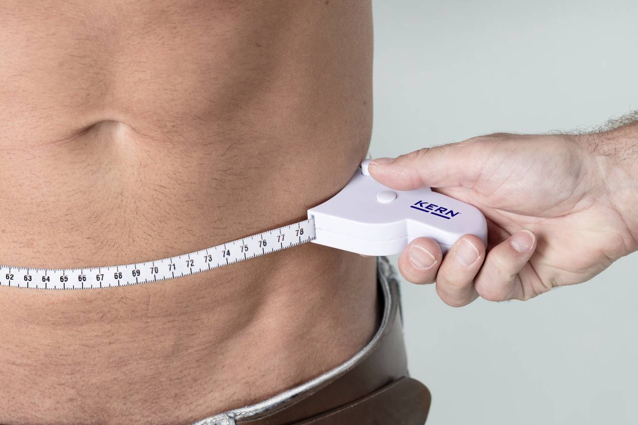 Ein Mann wird an der Taille gemessen. Eine Hand hält ein Maßband, das um seinen Bauch gelegt ist.