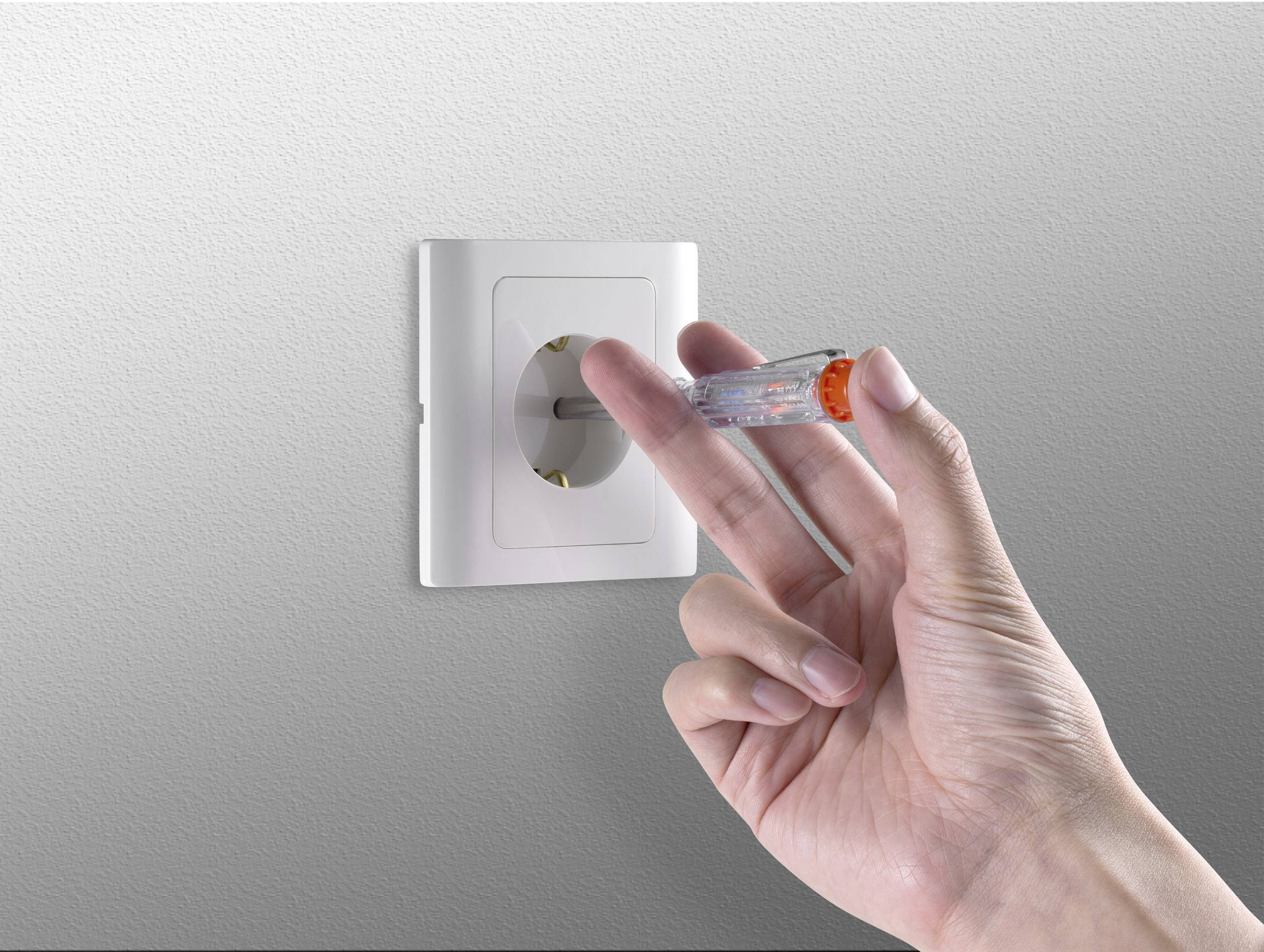 Eine Hand hält einen Spannungsprüfer nahe einer weißen Steckdose an einer grauen Wand, um elektrische Spannung zu überprüfen.