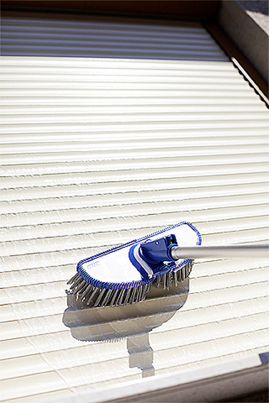 Ein Fensterladen wird mit einer Bürste gereinigt. Die Bürste hat weiße Borsten und einen blauen Griff. Das Sonnenlicht wirft Schatten.